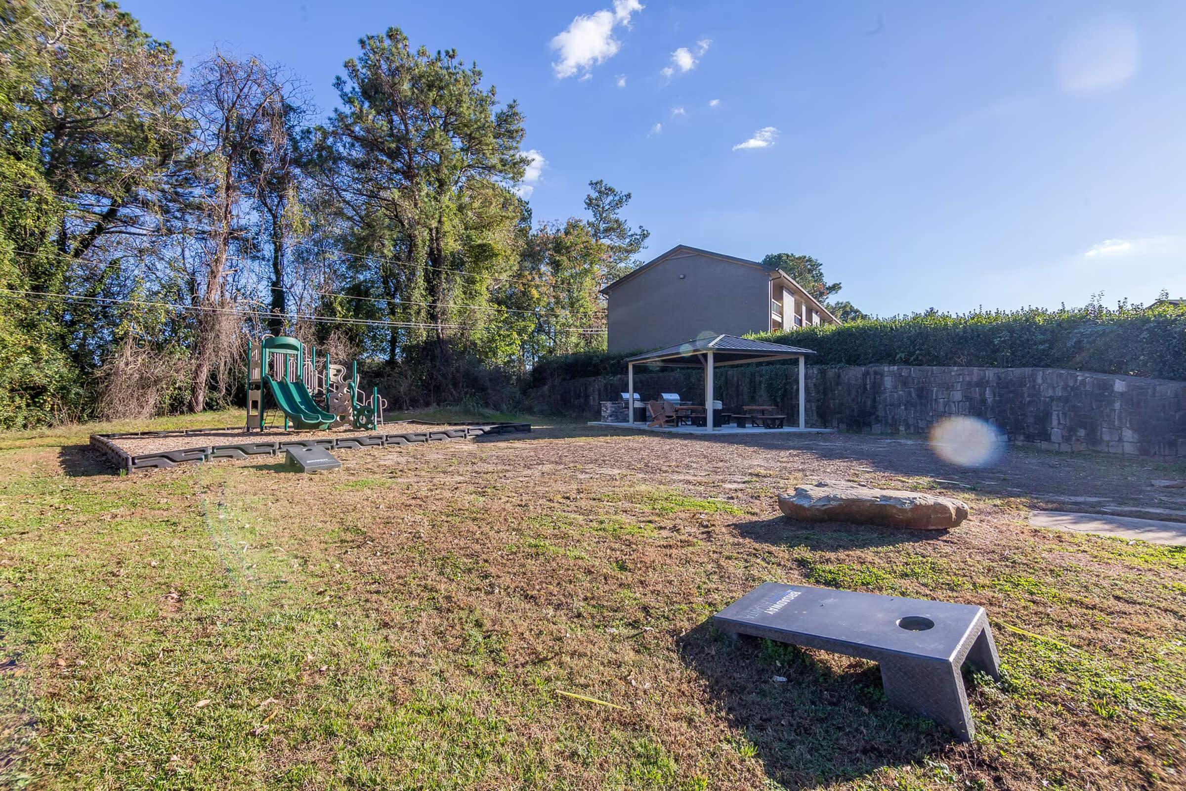 A playground area featuring a green slide and climbing structure surrounded by trees. There is a grassy space with benches and a picnic area under a shelter, and a simple game board on the ground. A building is visible in the background, under a clear blue sky.