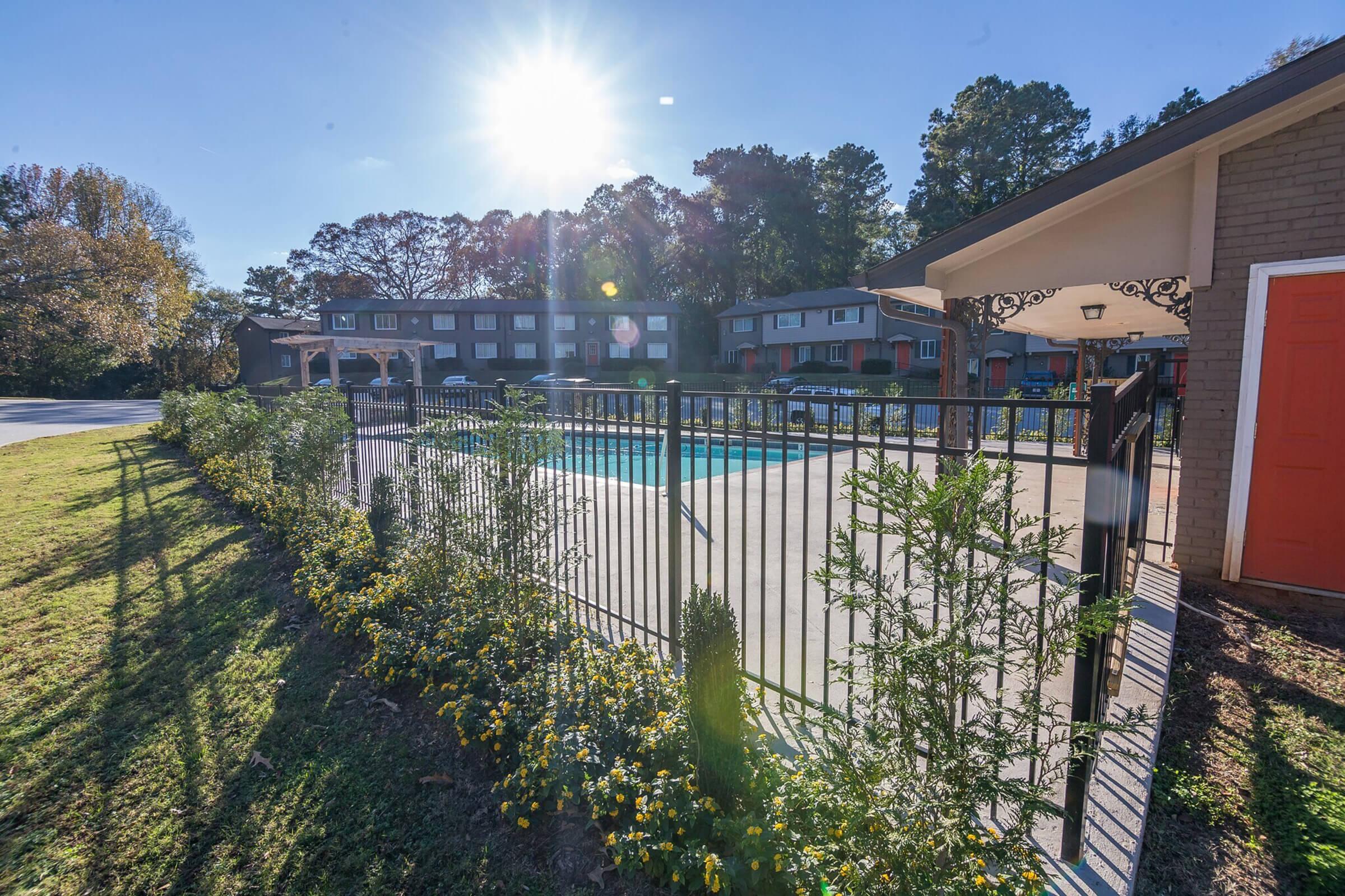 A well-maintained swimming pool area surrounded by a black metal fence and colorful flower beds, with apartment buildings in the background. The sun shines brightly in the clear blue sky, casting light on the scene.