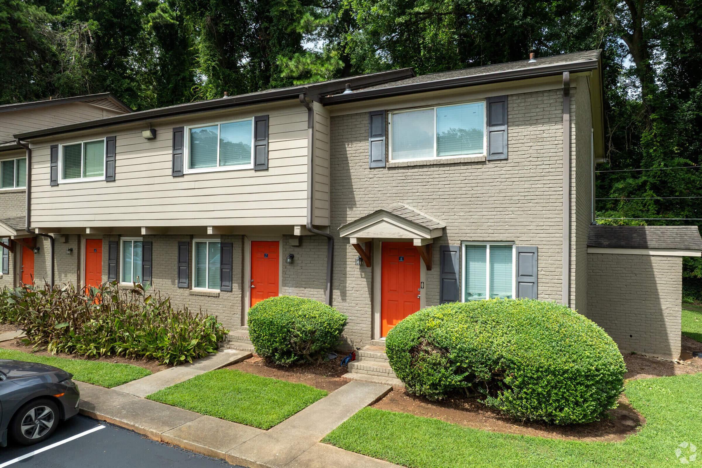 A two-story townhouse with gray brick exterior and tan siding. The front features orange doors and small windows. There is well-maintained landscaping including shrubs and grass in front, along with a parking area in the foreground. Trees are visible in the background.
