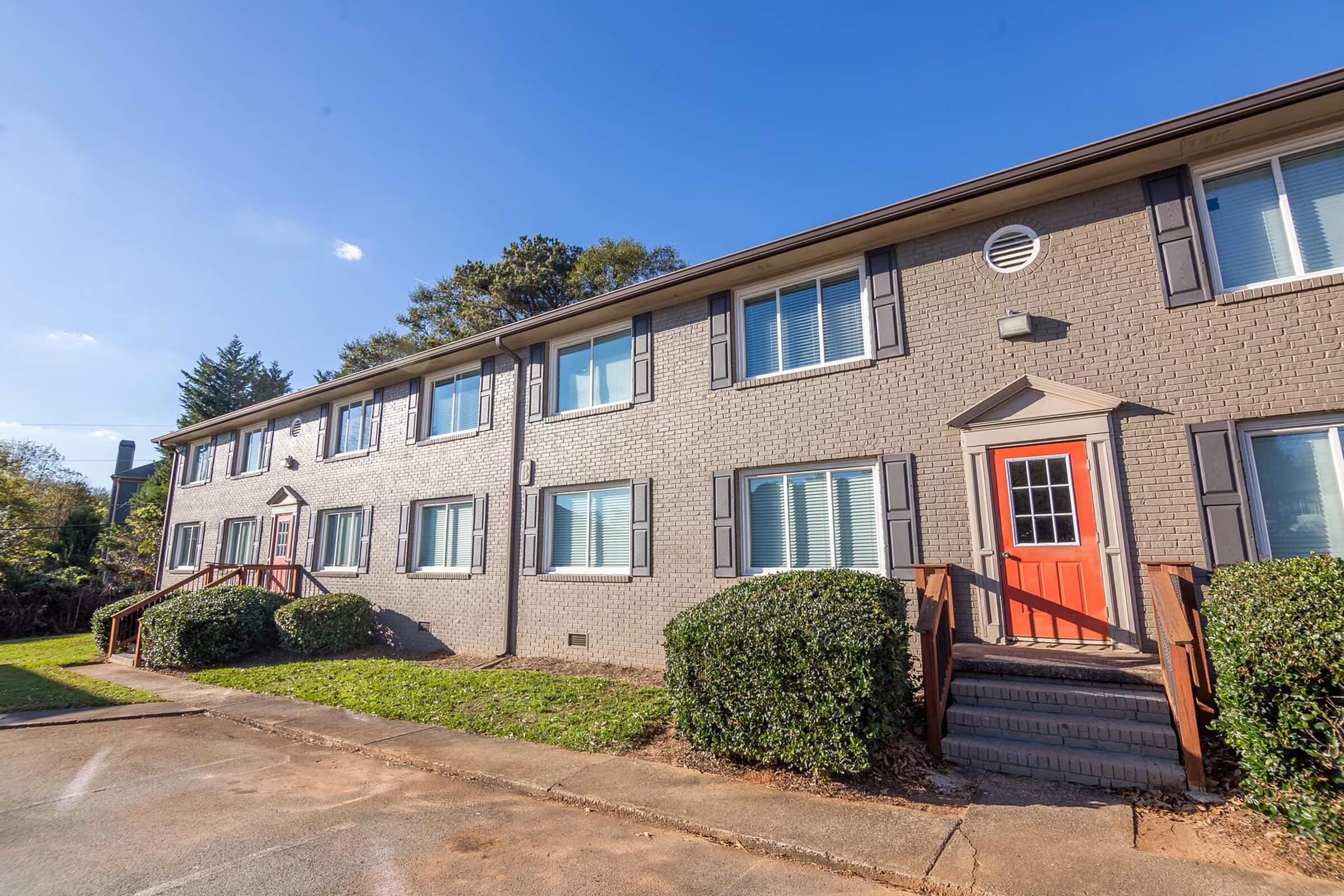 A brick apartment building with multiple windows and a distinctive orange door. The exterior features light gray brick, landscaped shrubs, and a paved pathway, under a clear blue sky. The building has a simple, modern design with a few steps leading to the entrance.