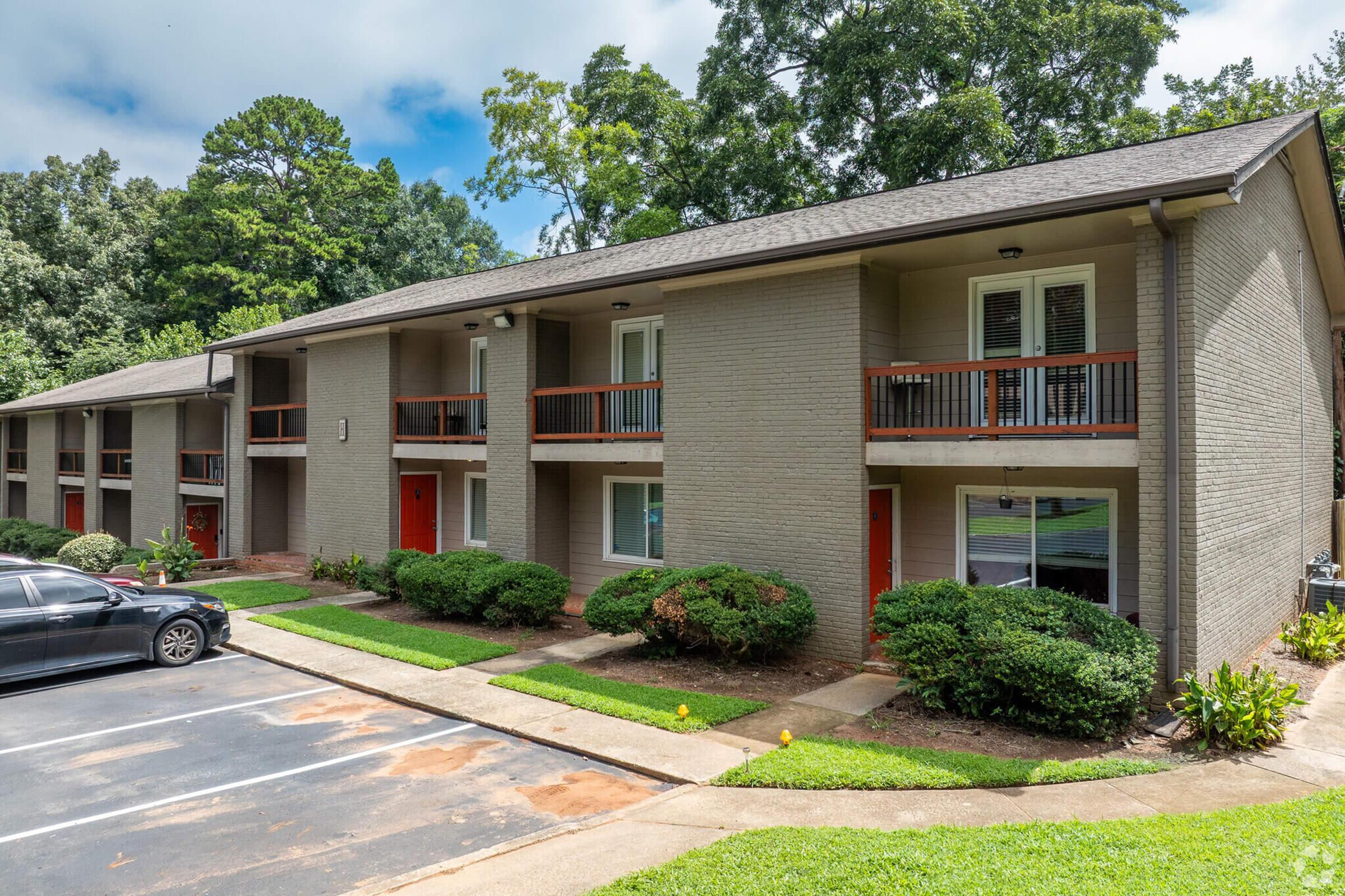 A view of a modern apartment building with two stories, featuring multiple units with red doors. The landscaped area has neatly trimmed bushes and a small parking lot in front. Trees and greenery surround the property, under a cloudy sky.