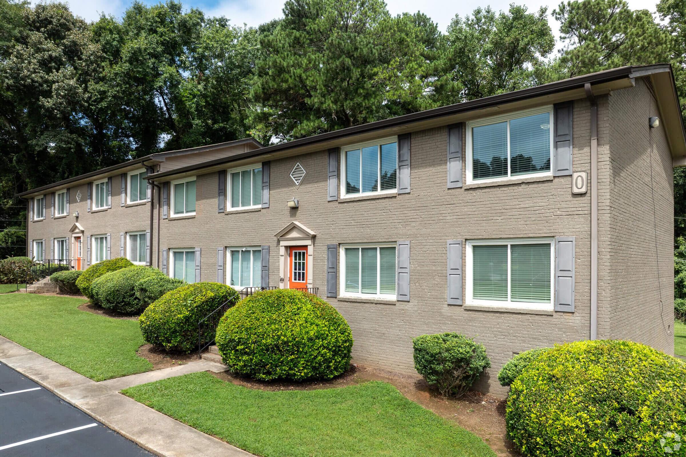 A two-story apartment building with a light gray brick exterior, featuring multiple windows and blue shutters. The building is surrounded by well-maintained green lawns and shrubs. A paved parking area is visible in the foreground, along with a tree line in the background.