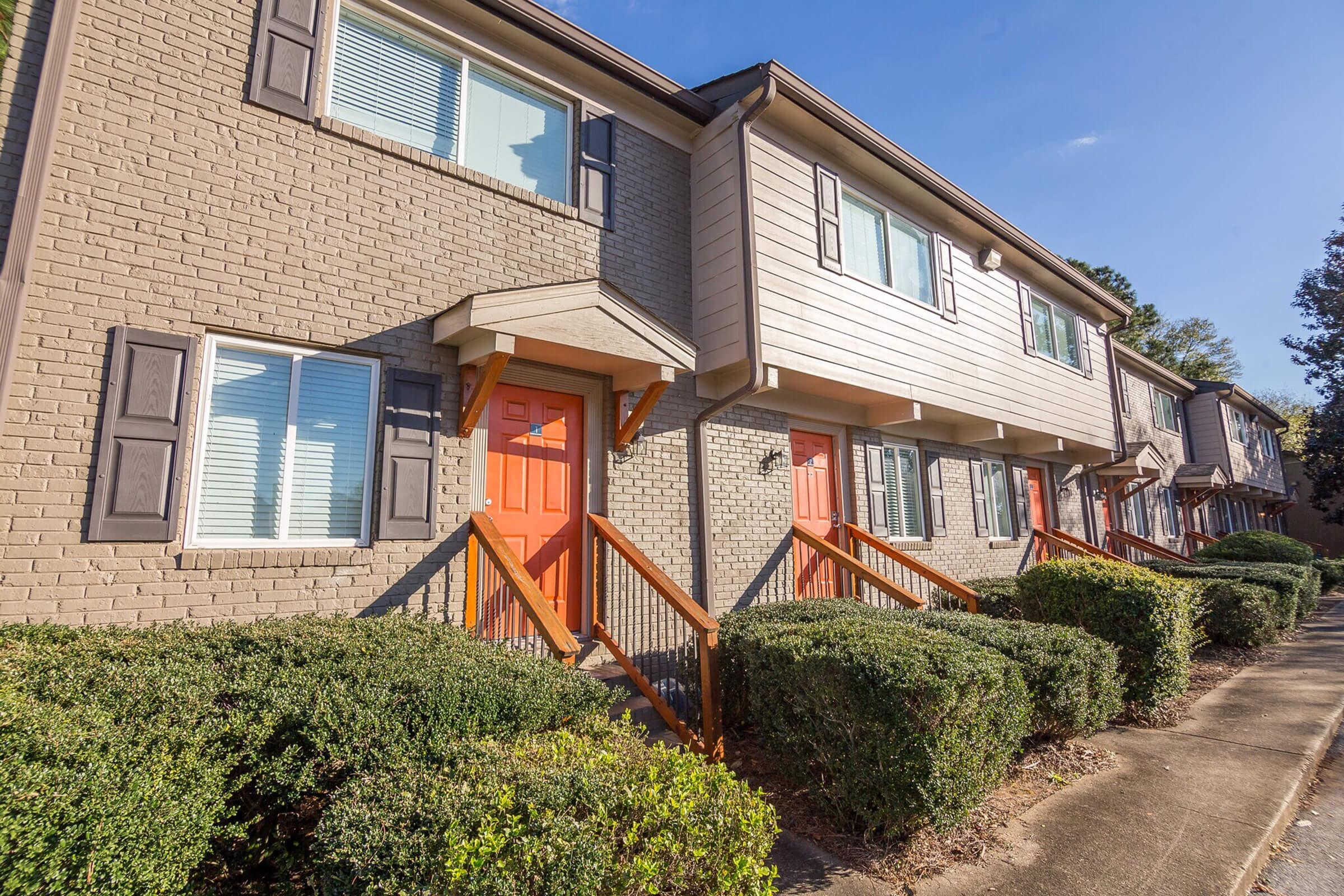 A row of two-story townhouses with a beige brick facade and orange doors. Each unit has windows with white frames and dark shutters. Neatly trimmed shrubs line the sidewalk in front of the homes, which are set against a clear blue sky.