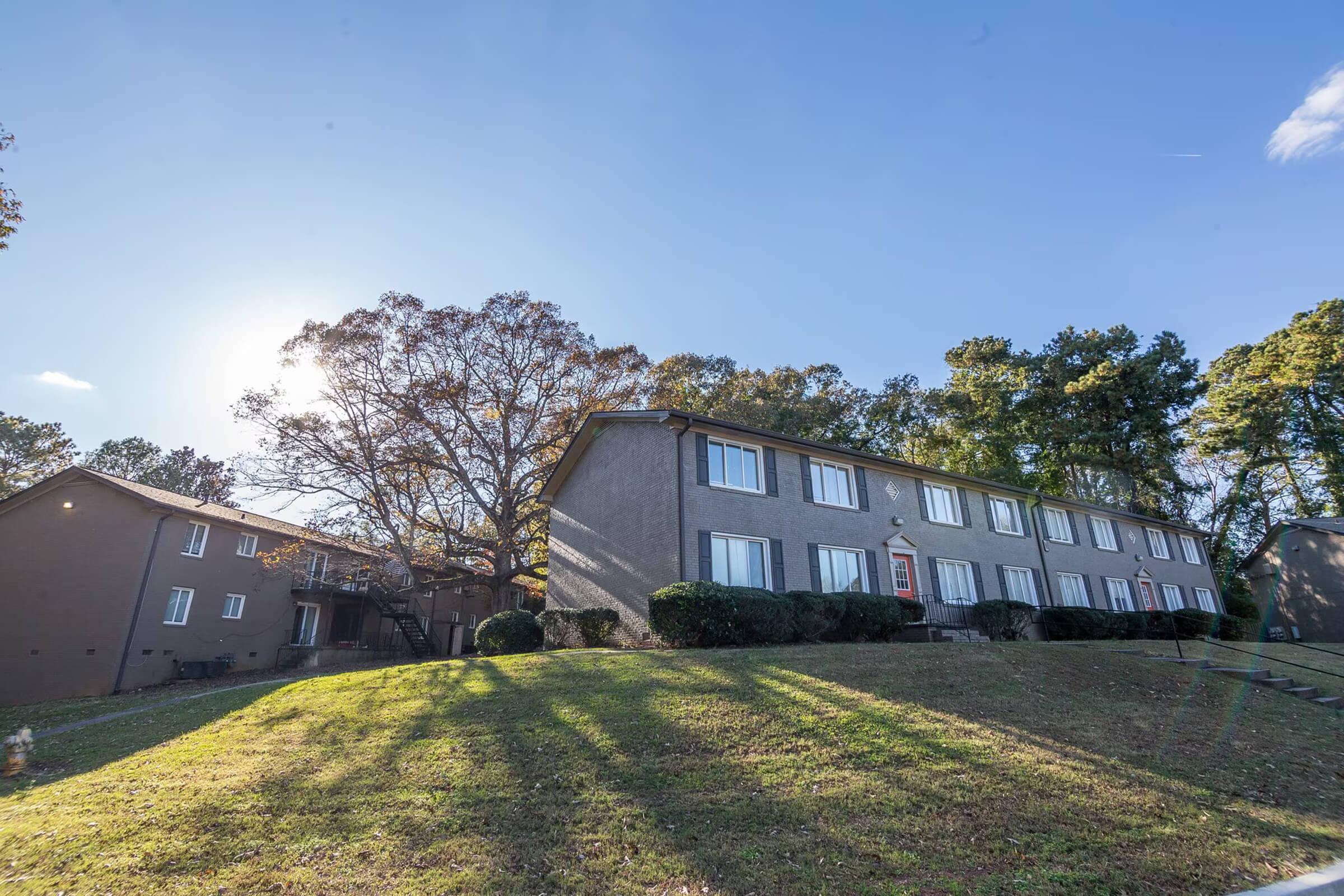 A multi-unit residential building surrounded by trees, situated on a grassy hill. The sun is shining in the background, casting long shadows from the trees onto the lawn. The building features multiple windows and a welcoming entrance. Another smaller building is visible to the left.