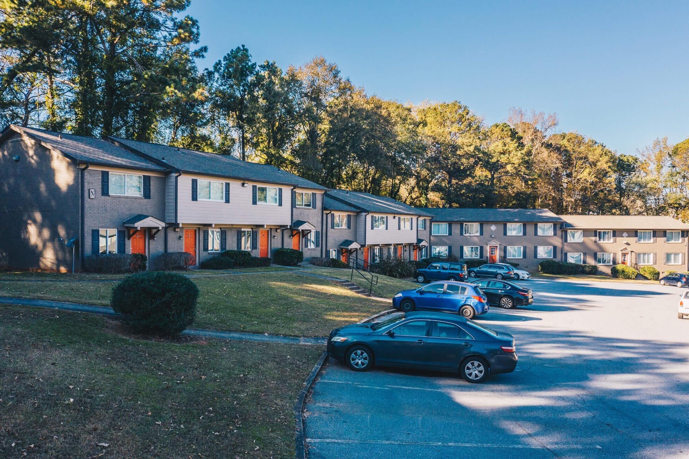 A residential area featuring two-story apartment buildings with a well-maintained lawn. Several parked cars line the driveway, and trees can be seen in the background under a clear blue sky. The buildings have a mix of neutral and contrasting colors, creating a welcoming neighborhood atmosphere.