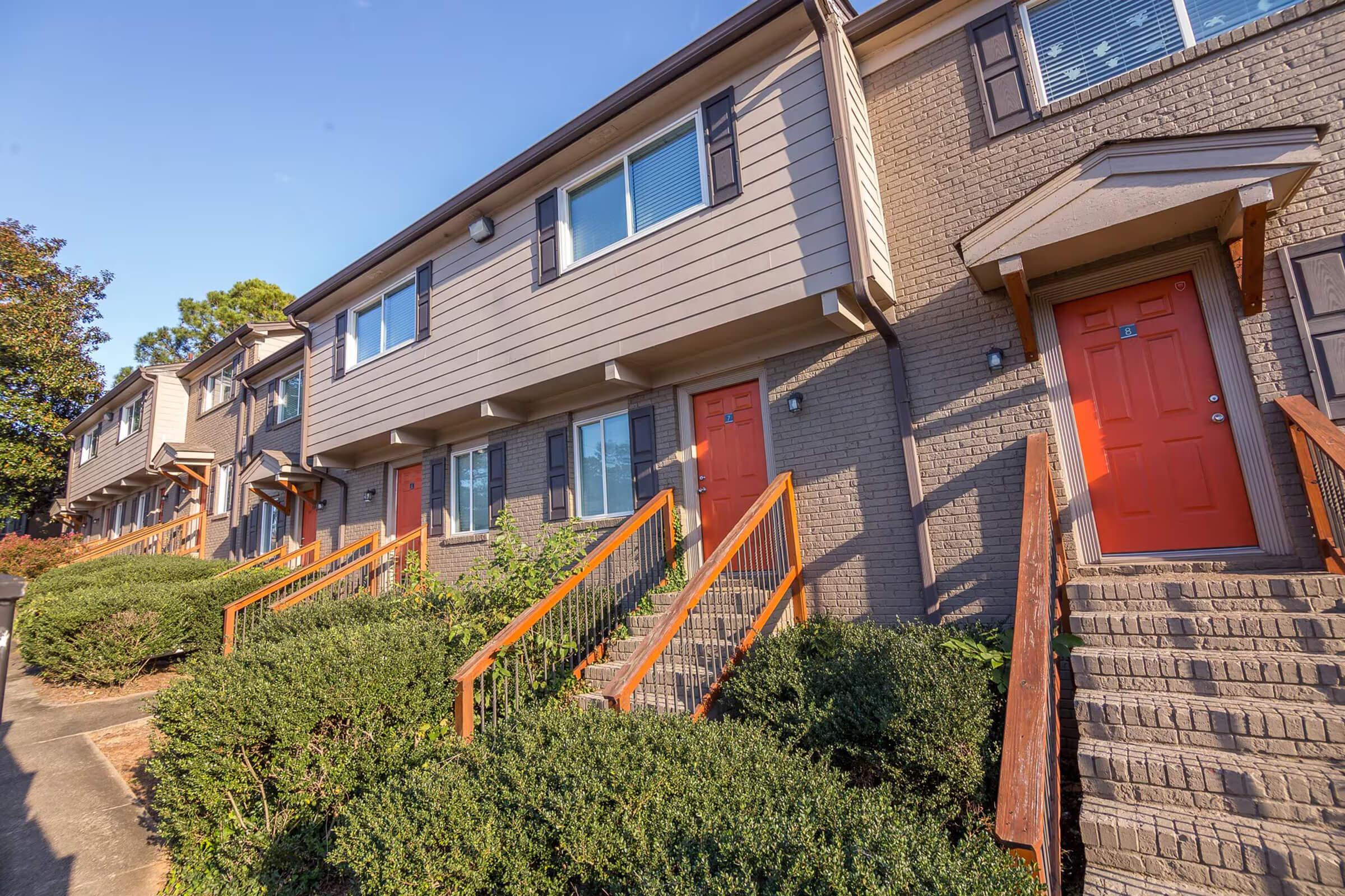 A row of modern townhouses featuring tan siding and bright orange doors. Each unit has a small staircase leading up to the entrance. The buildings are surrounded by neatly trimmed bushes and trees, under a clear blue sky. The setting conveys a welcoming residential atmosphere.