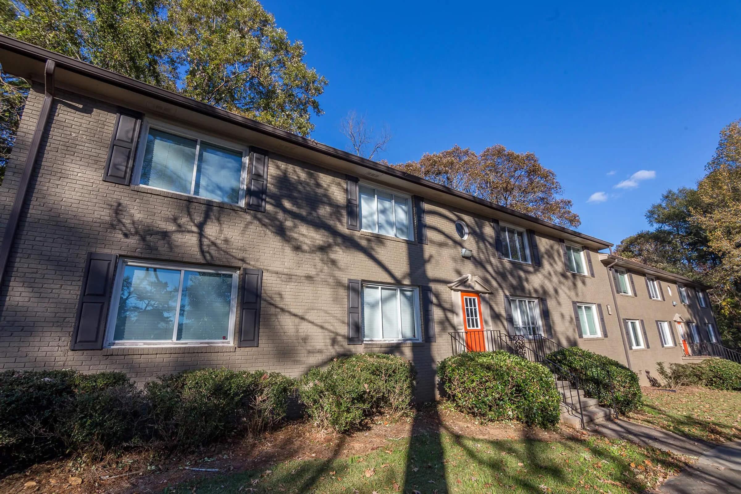 A two-story apartment building with a gray brick exterior. It features multiple windows, dark shutters, and a bright orange door. The surrounding area includes manicured bushes and trees, with blue sky and scattered clouds in the background. Shadows of trees are visible on the building's facade.