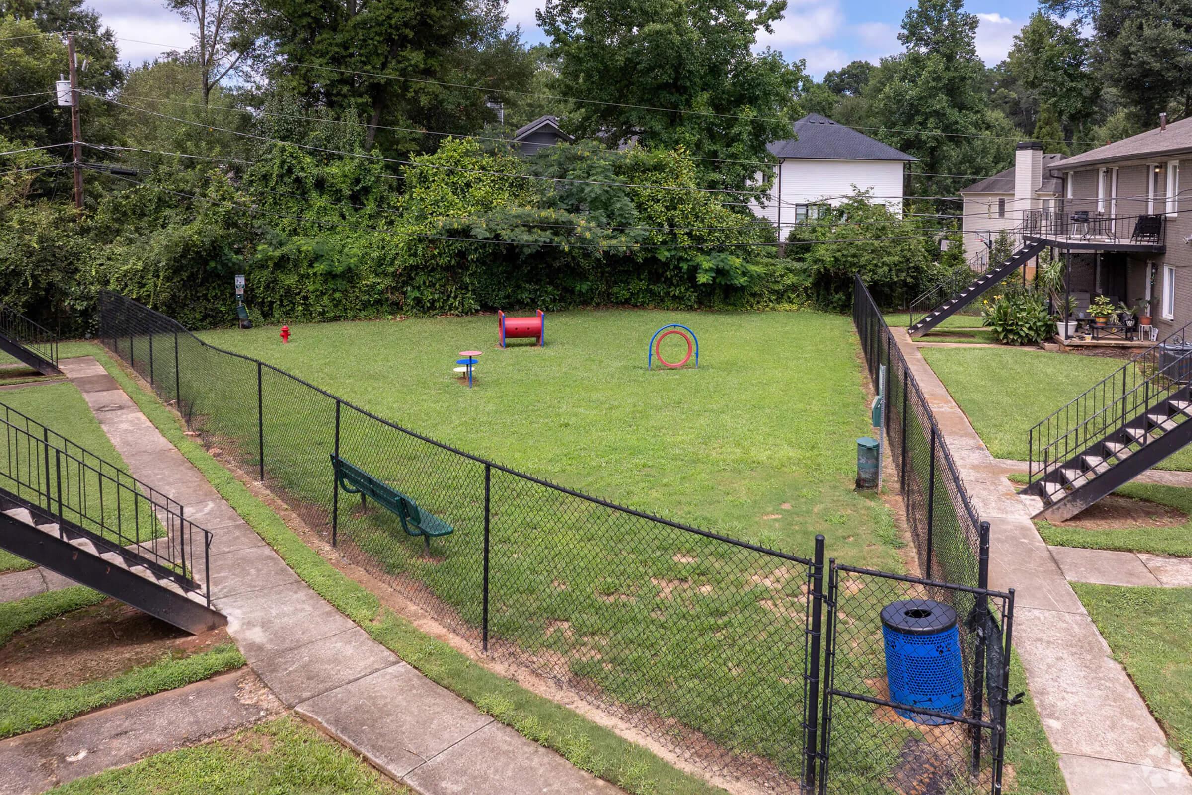 A fenced dog park featuring a grassy area with a few colorful play structures, including a tunnel and a small red house. Surrounding the park are pathways leading to adjacent buildings, and trees provide some shade in the background. The area is open and inviting for pets and their owners.