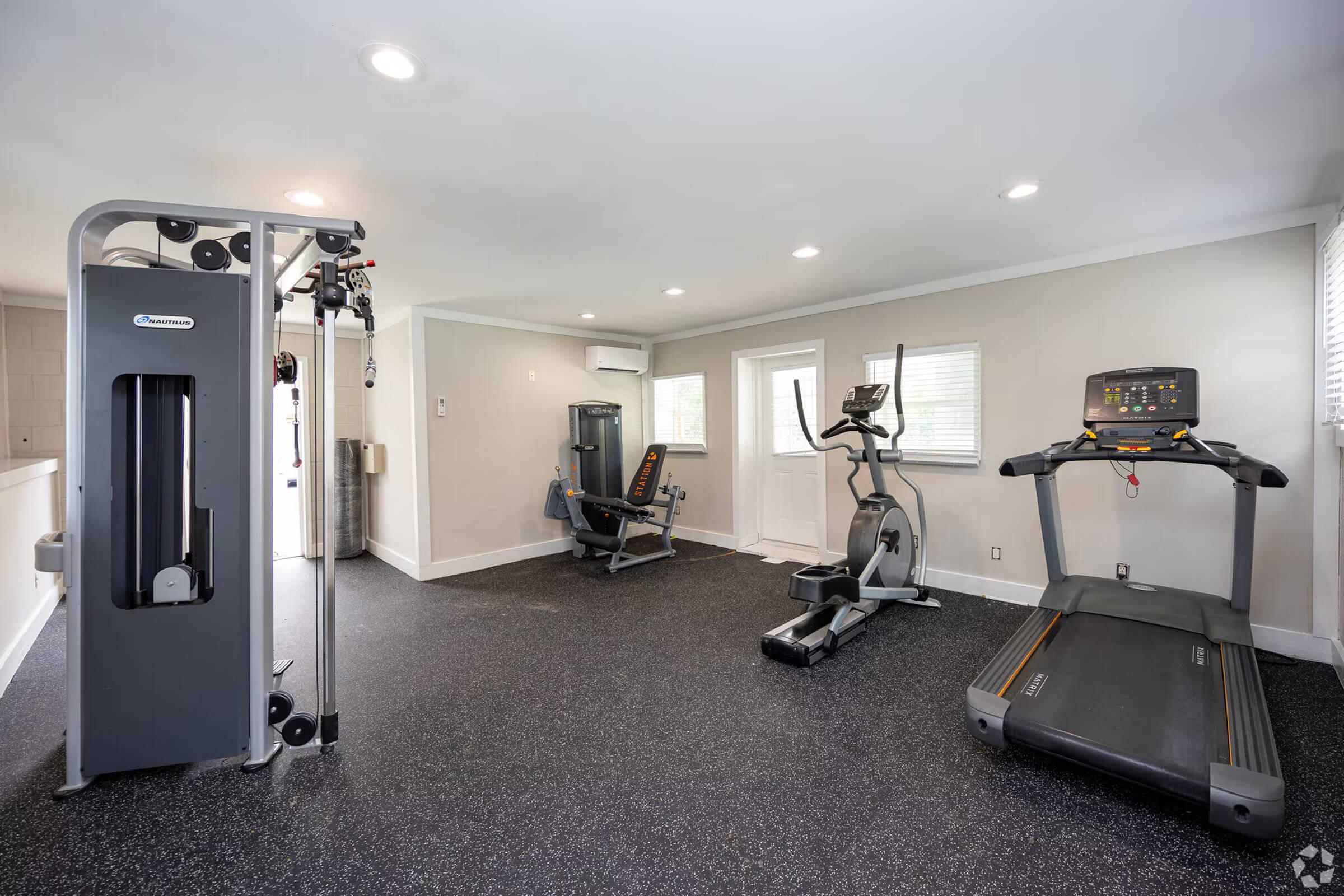 A well-lit home gym featuring a treadmill, elliptical machine, multi-gym weight station, and a workout mat. Large windows provide natural light, and the floor is covered with dark rubber flooring for durability and comfort.