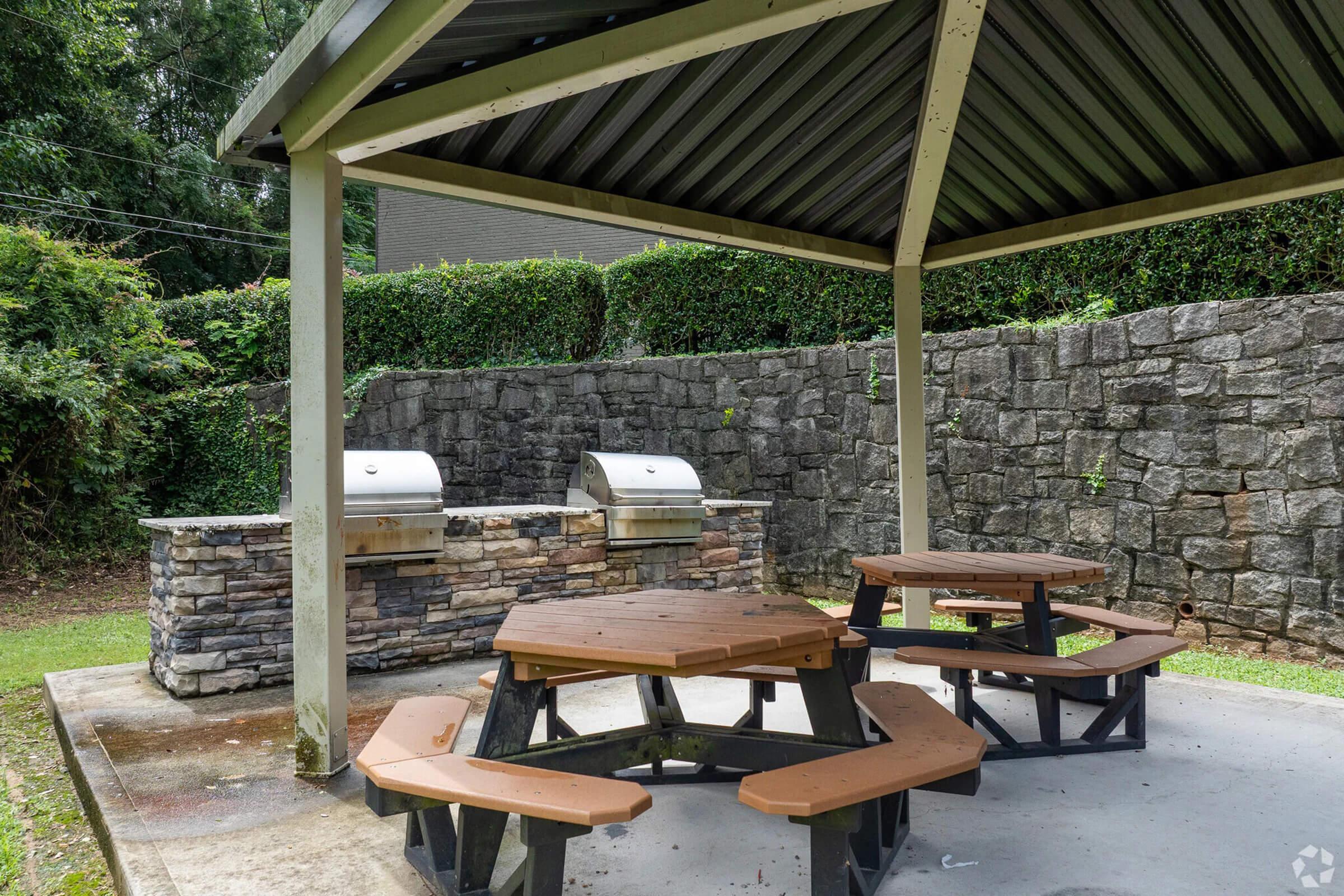 Outdoor barbecue area featuring a stone grill under a shaded pavilion. There are two metal grills and a picnic table made of segmented wooden benches arranged around a concrete surface. Lush greenery borders the space, with a stone wall in the background.