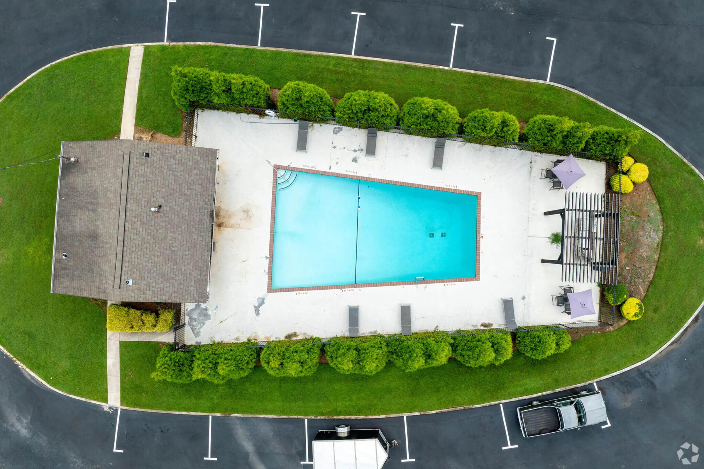 Aerial view of a rectangular swimming pool surrounded by green grass and hedges. The pool area includes lounge chairs and shaded seating under umbrellas, with a nearby building and parking spaces visible. The scene is well-maintained and appears inviting for recreational use.