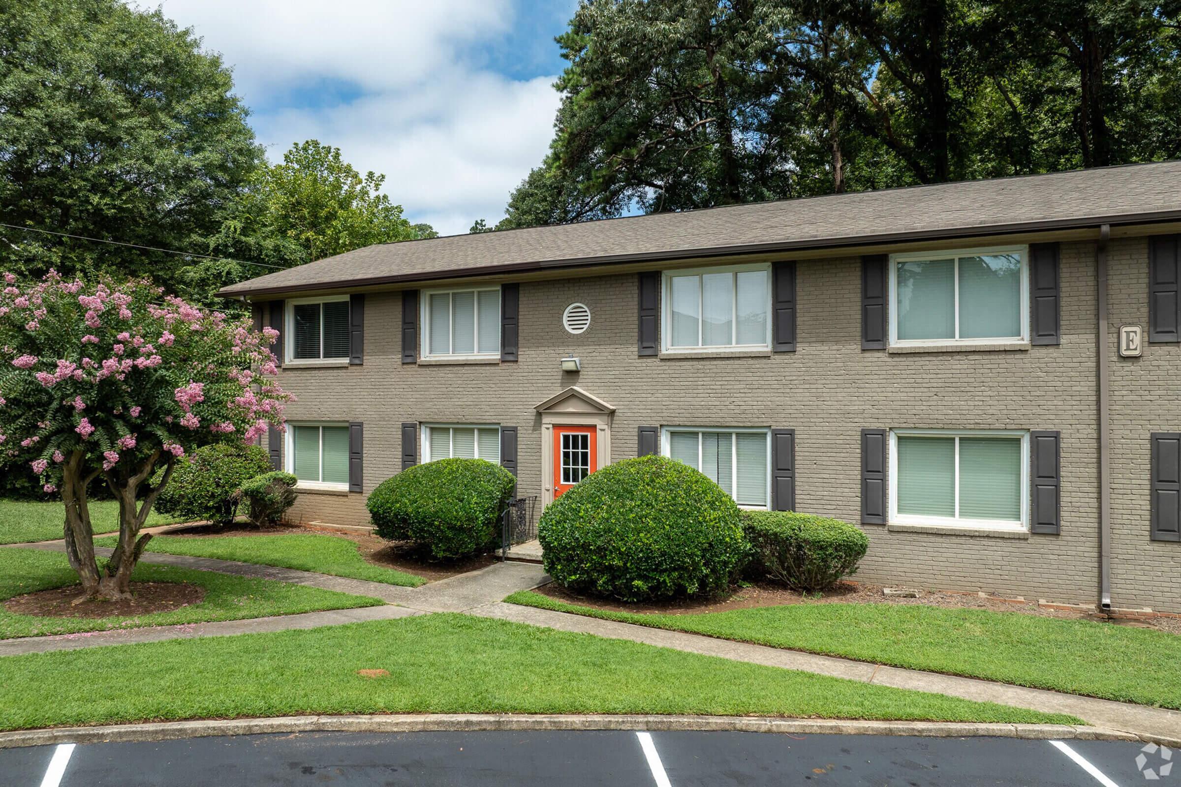 Two-story residential building with gray brick exterior, featuring multiple windows with shutters. In front, there are neatly trimmed shrubs and a flowering tree. A pathway leads to the orange front door. The surrounding lawn is green and well-maintained, with a cloudy sky overhead.