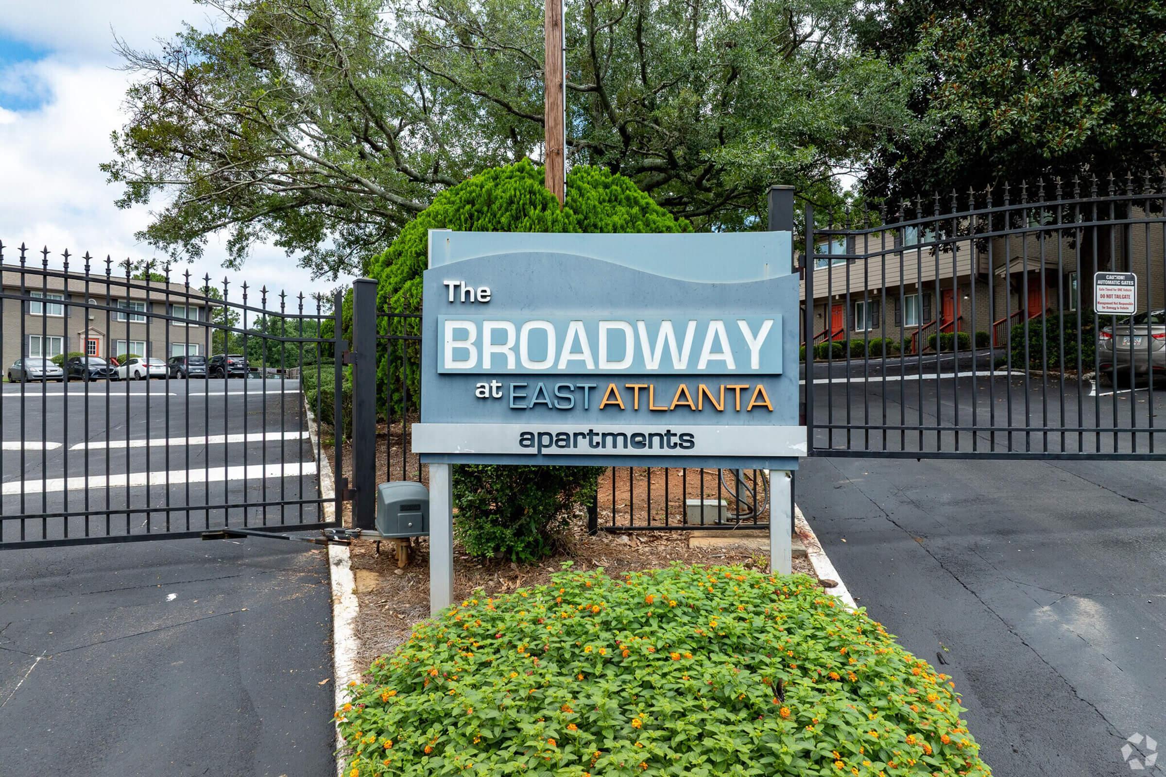 Sign for "The Broadway at East Atlanta Apartments," displaying the name prominently. The sign is located near a gated entrance with greenery and a well-maintained landscape. The background features apartment buildings, and the scene is set on a partly cloudy day. 