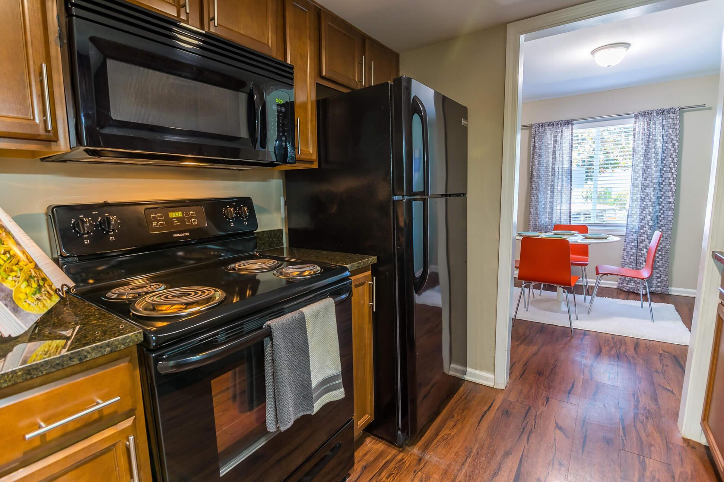 A modern kitchen featuring dark wood cabinets, a black stove and refrigerator, and a granite countertop. A window lets in natural light, illuminating the nearby dining area with orange chairs and a small table. The flooring is a warm wood laminate, enhancing the inviting atmosphere.
