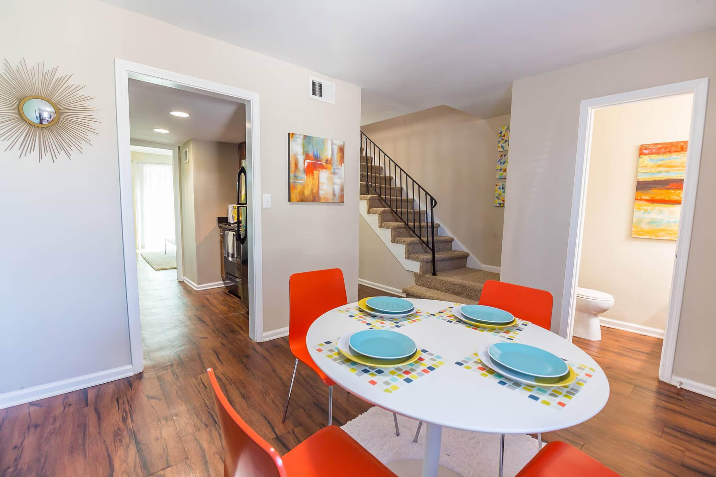 A bright and modern dining area featuring a round white table set with blue plates, surrounded by orange chairs. The walls are painted in neutral tones, and there's a stylish sunburst wall clock. In the background, a staircase leads to the upper floor, and an inviting hallway is visible.