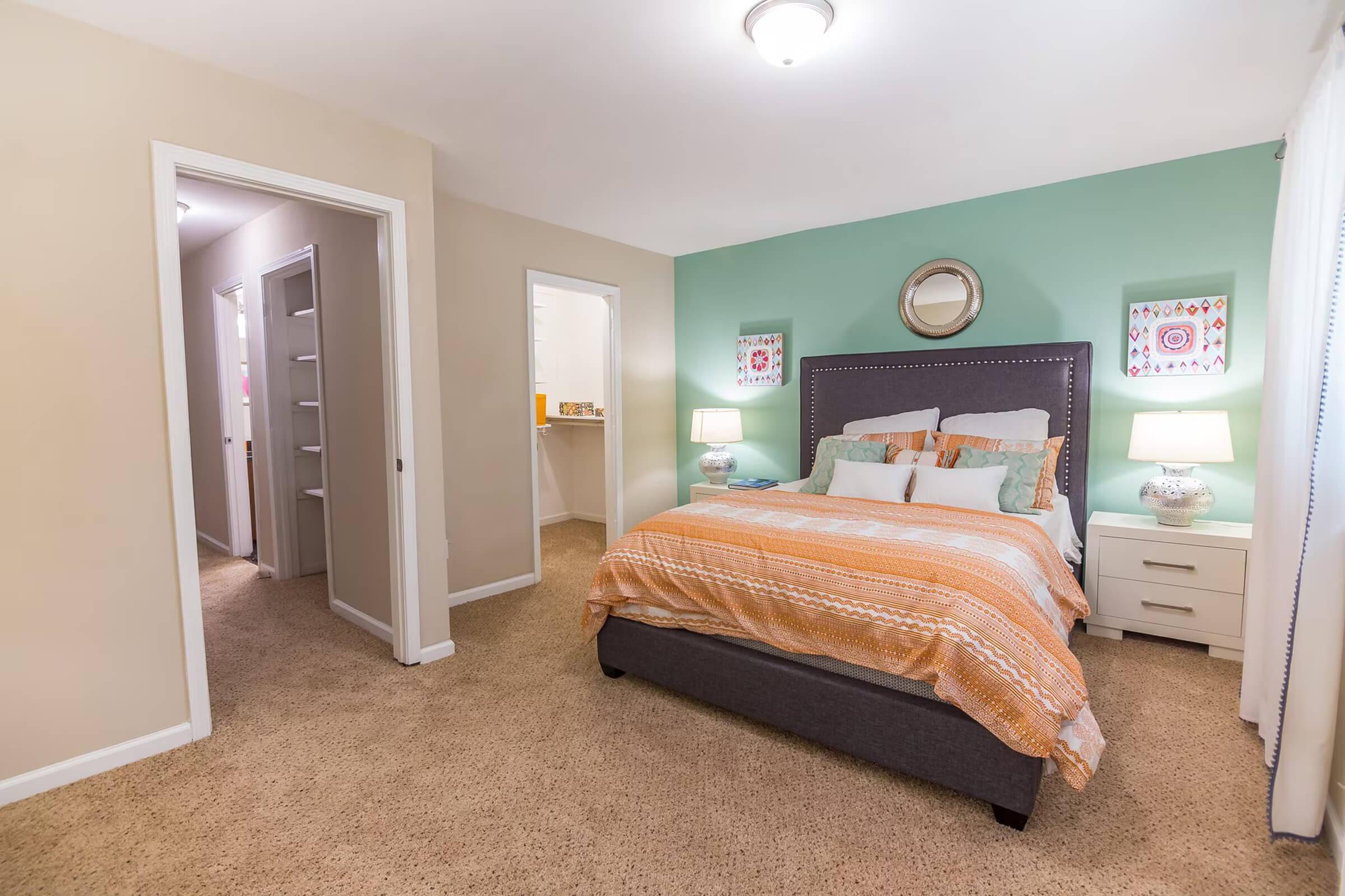 A cozy bedroom featuring a queen-sized bed with orange and white bedding, two bedside tables with lamps, and light green accent walls. A doorway leads to a hallway, and another door opens to a small room, possibly a bathroom or closet. The carpeted floor adds warmth to the space.