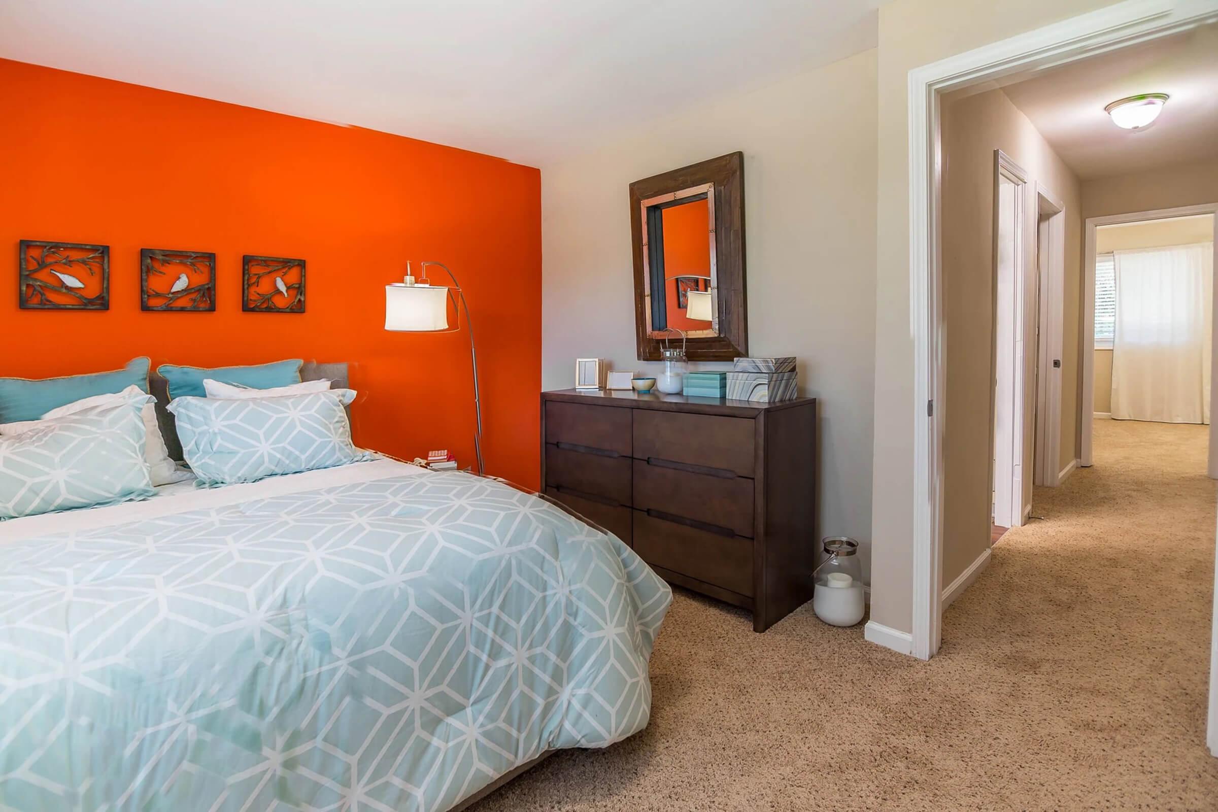 Brightly colored bedroom featuring an orange accent wall, a cozy bed with light blue and white bedding, and decorative wall art. A wooden dresser is positioned beside the bed, and a mirror hangs above it. The room has a soft carpet and leads to a well-lit hallway with doors on the right.
