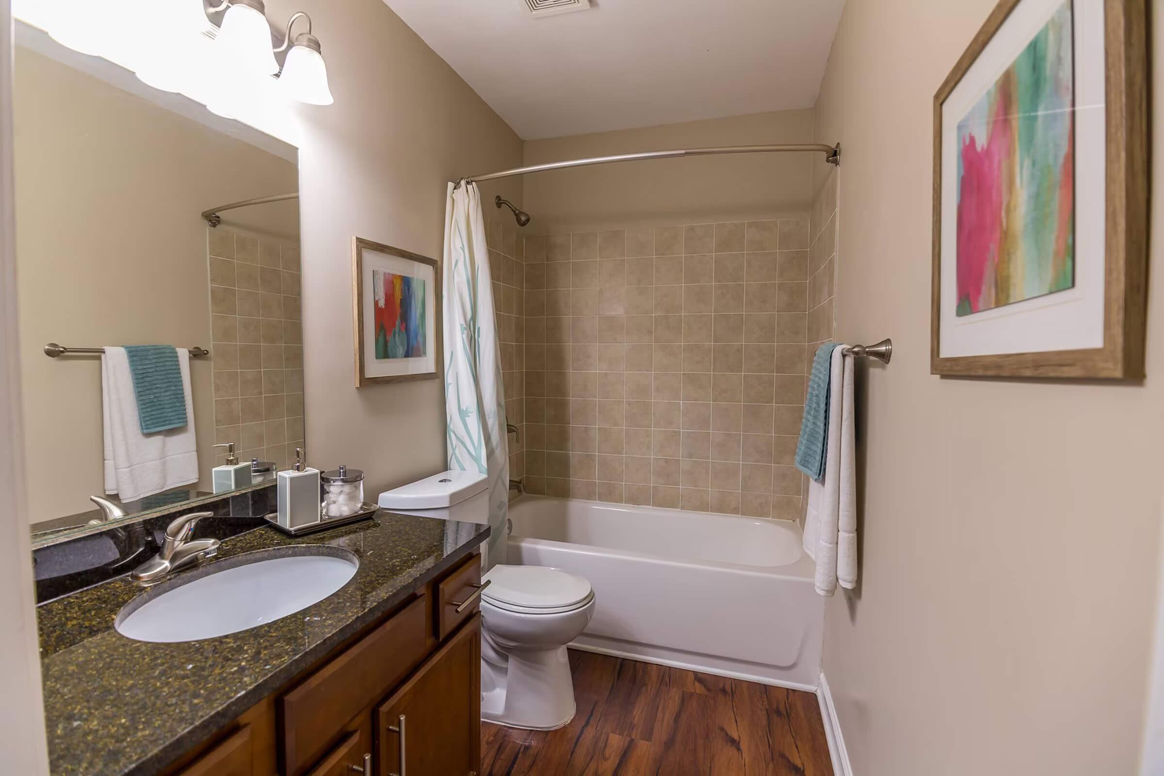 A well-lit bathroom featuring a shower-tub combo with a curtain, a sink with a dark granite countertop, and a toilet. Towels hang on a rack, and there's a framed colorful art piece on the wall. The floor has wood-like tiles, creating a warm atmosphere.