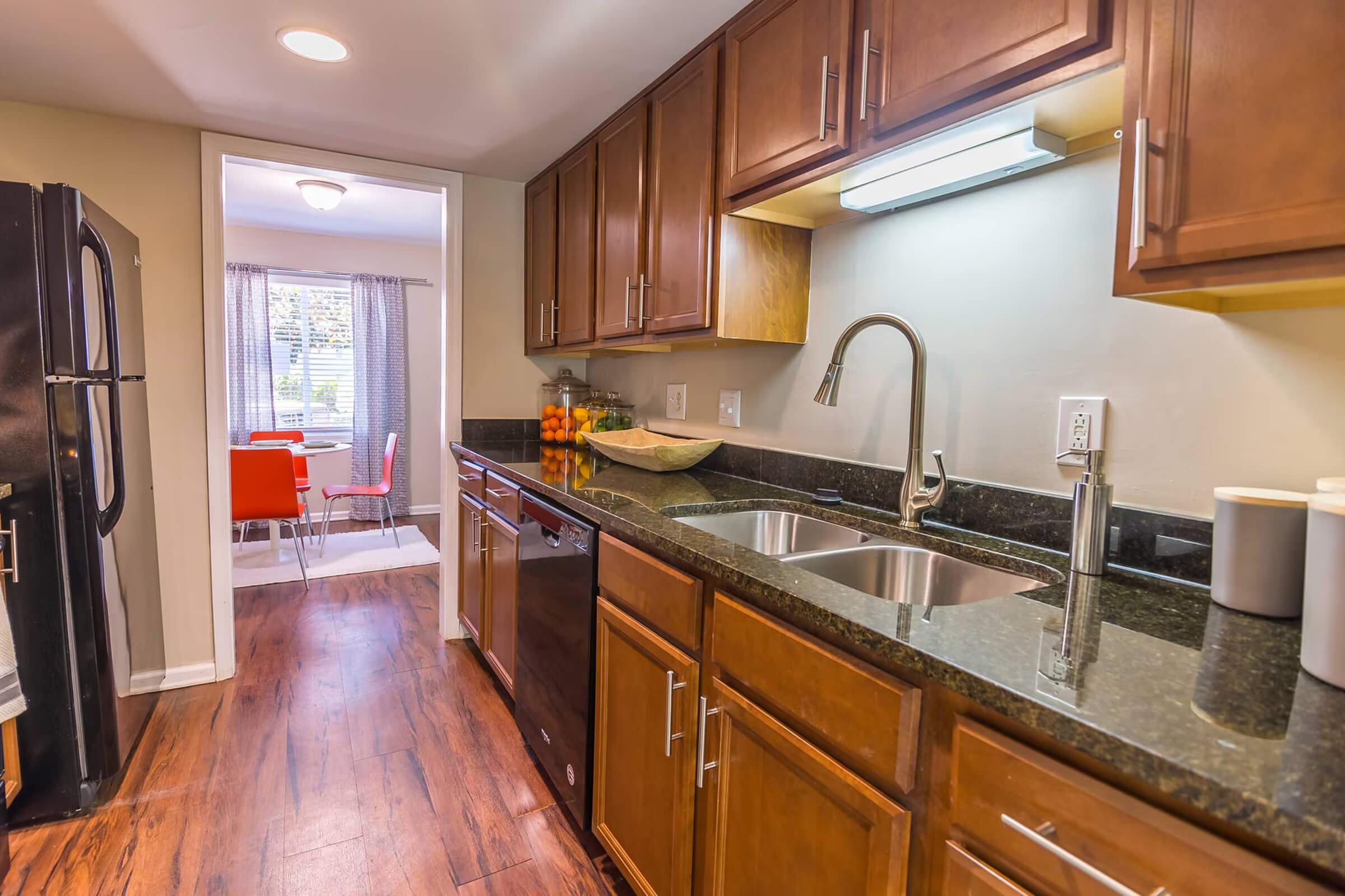 A modern kitchen featuring wooden cabinets, a double sink, and a dark granite countertop. Stainless steel appliances, including a dishwasher and refrigerator, are visible. The space transitions to a dining area with bright red chairs and a table, illuminated by natural light from a nearby window.