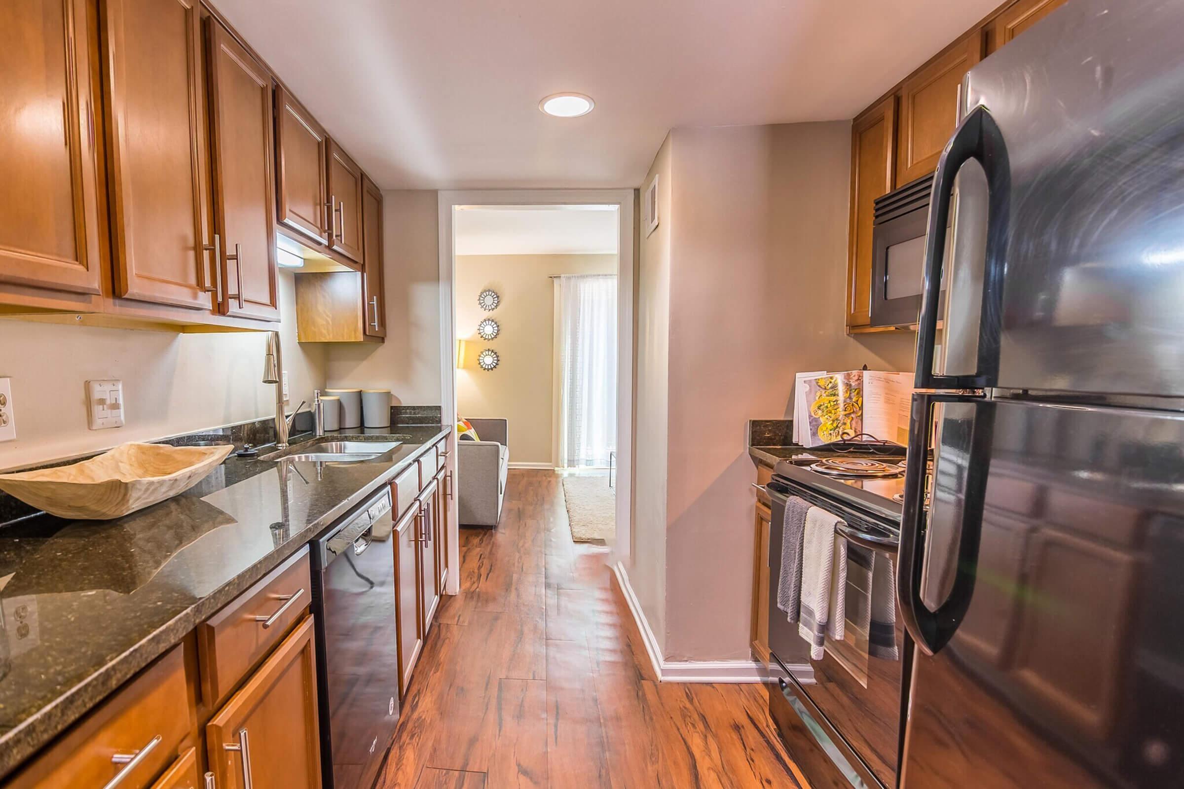 Modern kitchen with wooden cabinets and dark countertops, featuring a stainless steel refrigerator and oven. A doorway leads to a bright living area with light-colored walls and decorative wall art. Natural light enters from the window, illuminating the space.