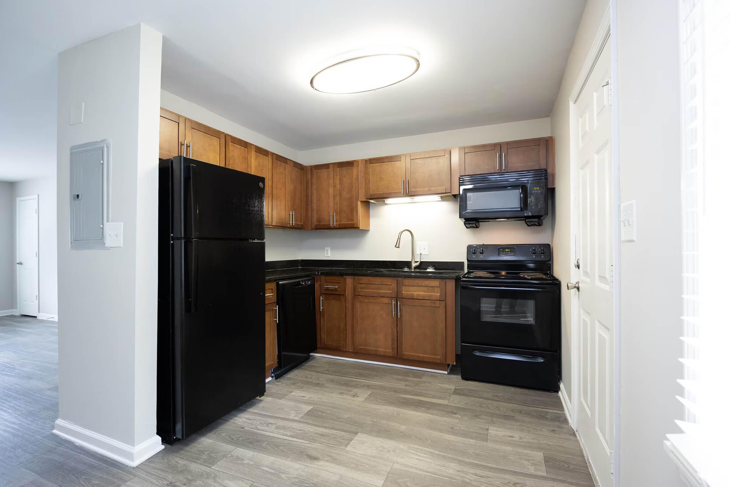 Modern kitchen featuring dark wood cabinets, black appliances including a refrigerator and stove, and a sleek countertop. The room is well-lit with a circular ceiling light and has a neutral color scheme, complemented by laminate flooring. A door leads to another area of the home.