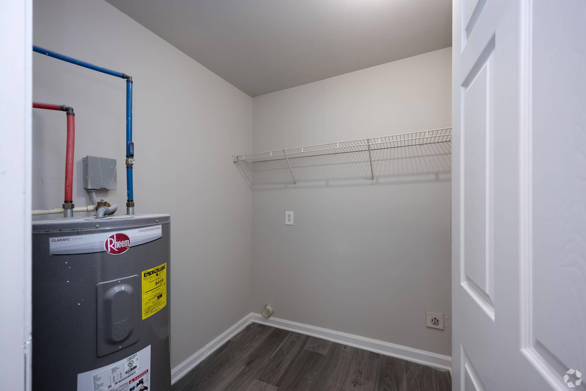 A small laundry room featuring a water heater with a yellow label, pipes, and a wall-mounted shelf. The walls are light-colored, and the floor is a dark laminate. The space is empty, with no appliances or furniture present.