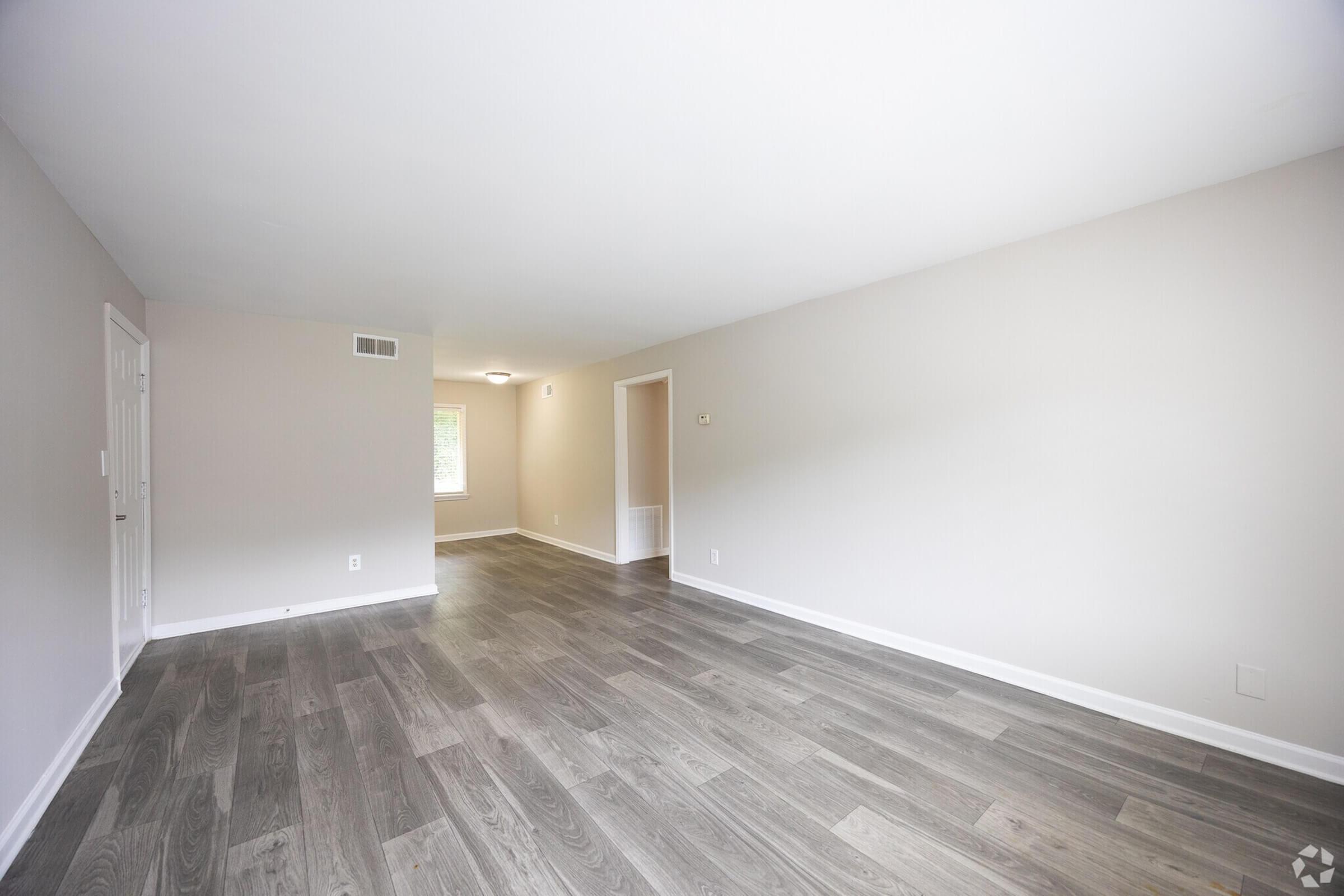 Spacious, empty living room with light-colored walls and gray wood flooring. Soft natural light enters through windows, creating a bright atmosphere. The layout includes a door on the left, leading to another room, and a light fixture on the ceiling. Ideal for various furniture arrangements.