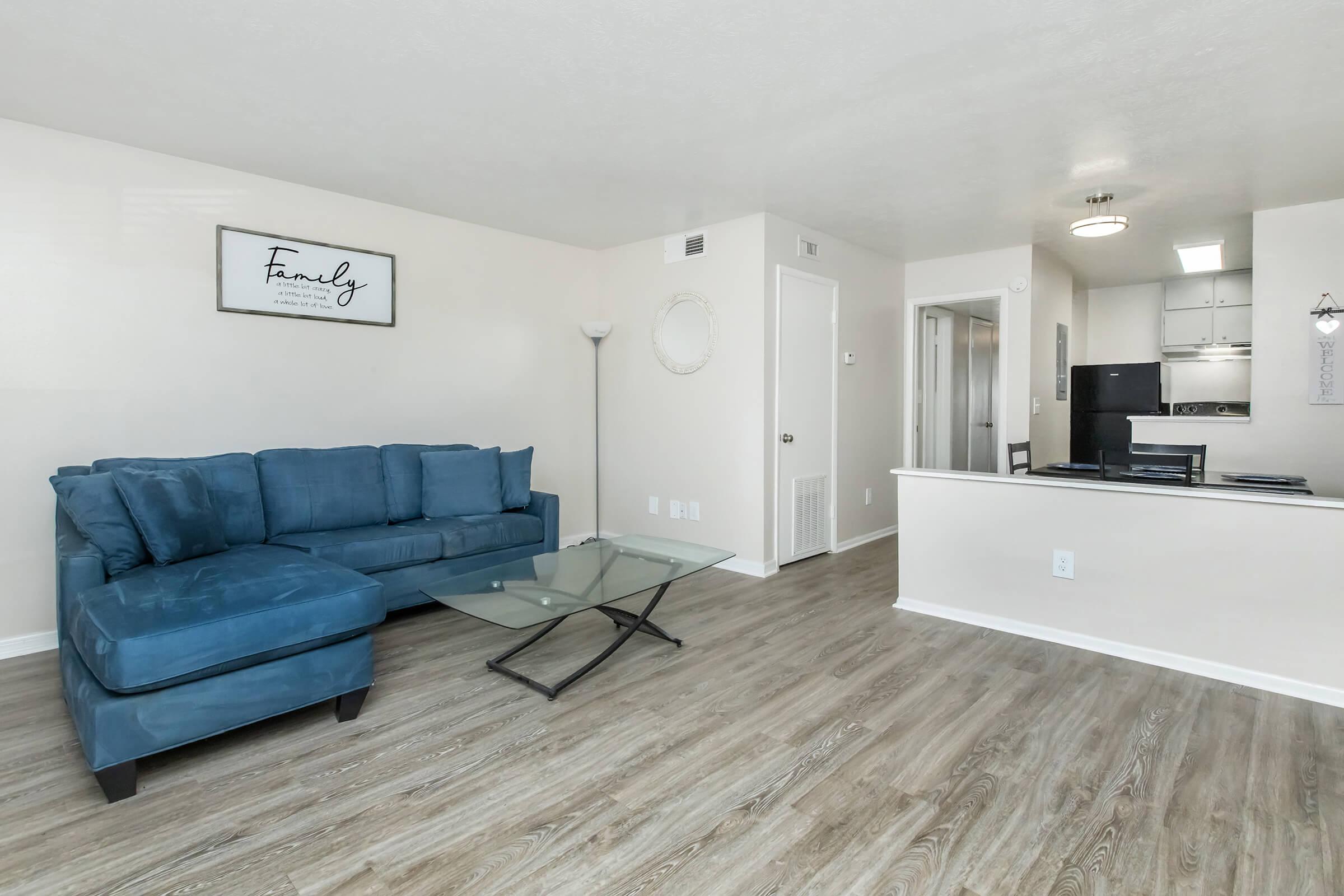 A modern living room featuring a blue sectional sofa, a glass coffee table, and light-colored walls. There is a wall decoration that says "Family" and an open-concept layout leading to a kitchen area. The flooring is a warm wood-like finish, creating a cozy atmosphere.