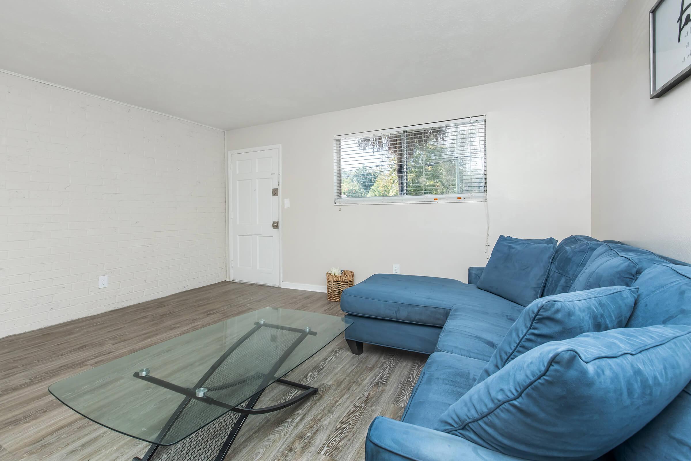 A bright living room featuring a large blue sectional sofa, a modern glass coffee table, and light-colored walls. A door is visible on the left, while a window allows natural light to enter the space. The flooring is wood-like, adding to the contemporary feel of the room.