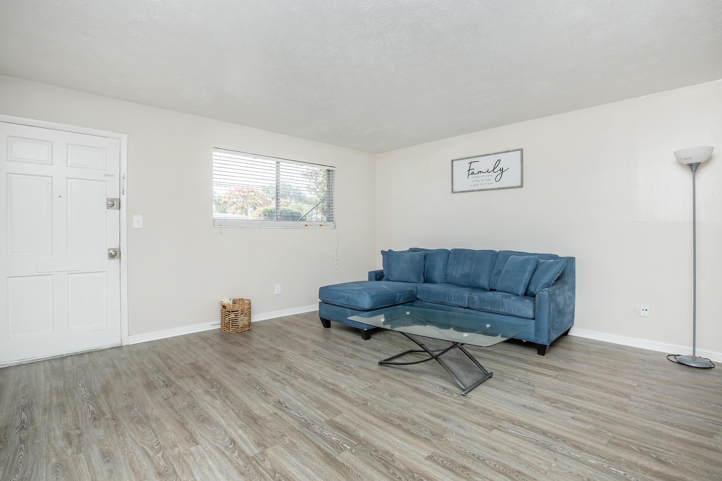 A small living room featuring a light blue sectional sofa, a glass coffee table, and a floor lamp. The walls are painted light beige, and there is a window with blinds allowing natural light. A decorative basket is placed near the entrance, and a framed sign with the word "Family" hangs on the wall.