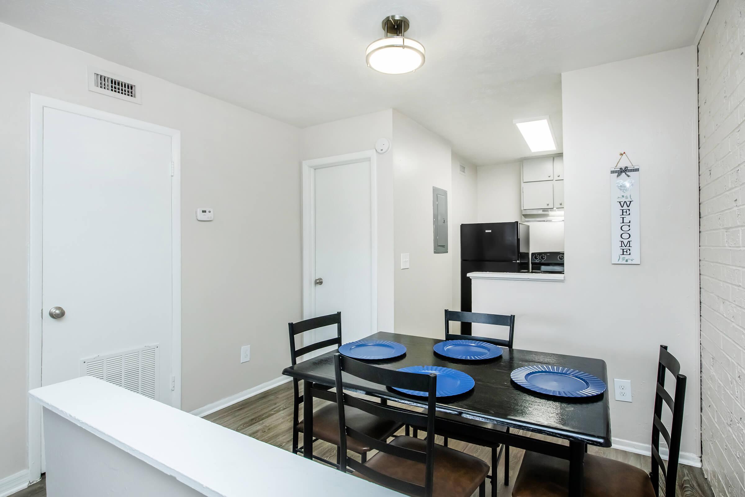 A modern kitchen area with a dining table set for four. The table is adorned with blue plates, and there are chairs around it. In the background, a kitchen is visible with white cabinetry and black appliances. The walls are a light color, and a small sign hangs nearby, welcoming guests.