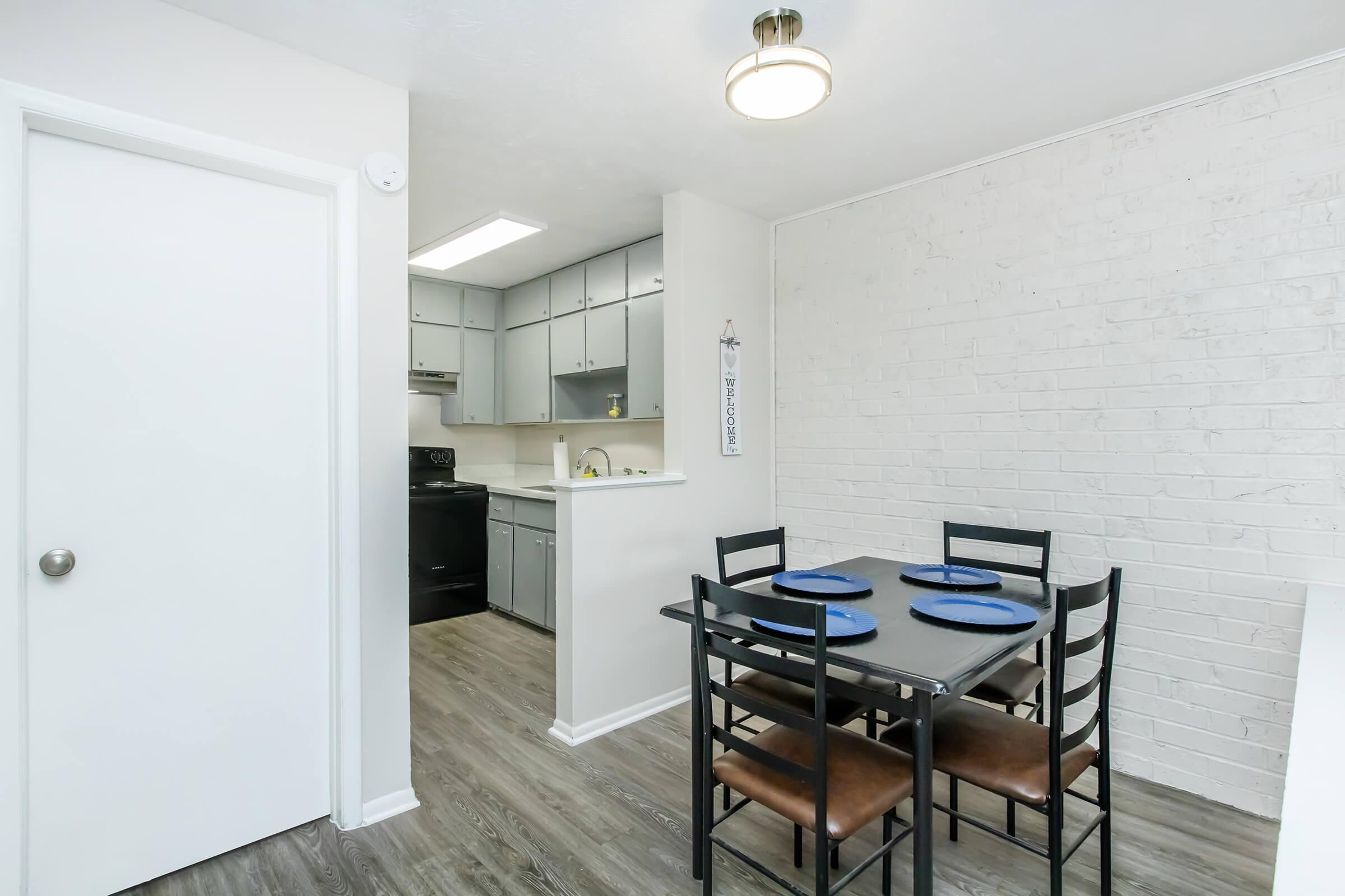 A modern dining area featuring a black table set with blue plates and four chairs. The space has white brick walls and a door leading to another room. A bright kitchen is visible in the background with grey cabinets and appliances. Ceiling light fixture adds warmth to the room.