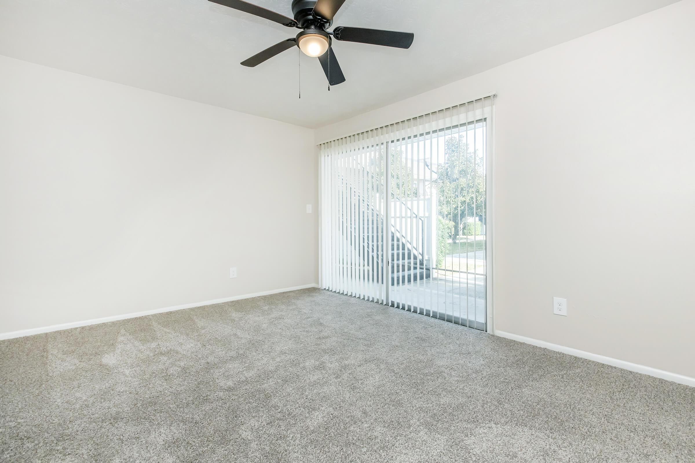 A spacious, empty room with light beige walls and plush gray carpet. A ceiling fan is mounted overhead. The room features a large sliding glass door with vertical blinds, leading to an outdoor area with visible stairs. Natural light brightens the space, creating a welcoming atmosphere.