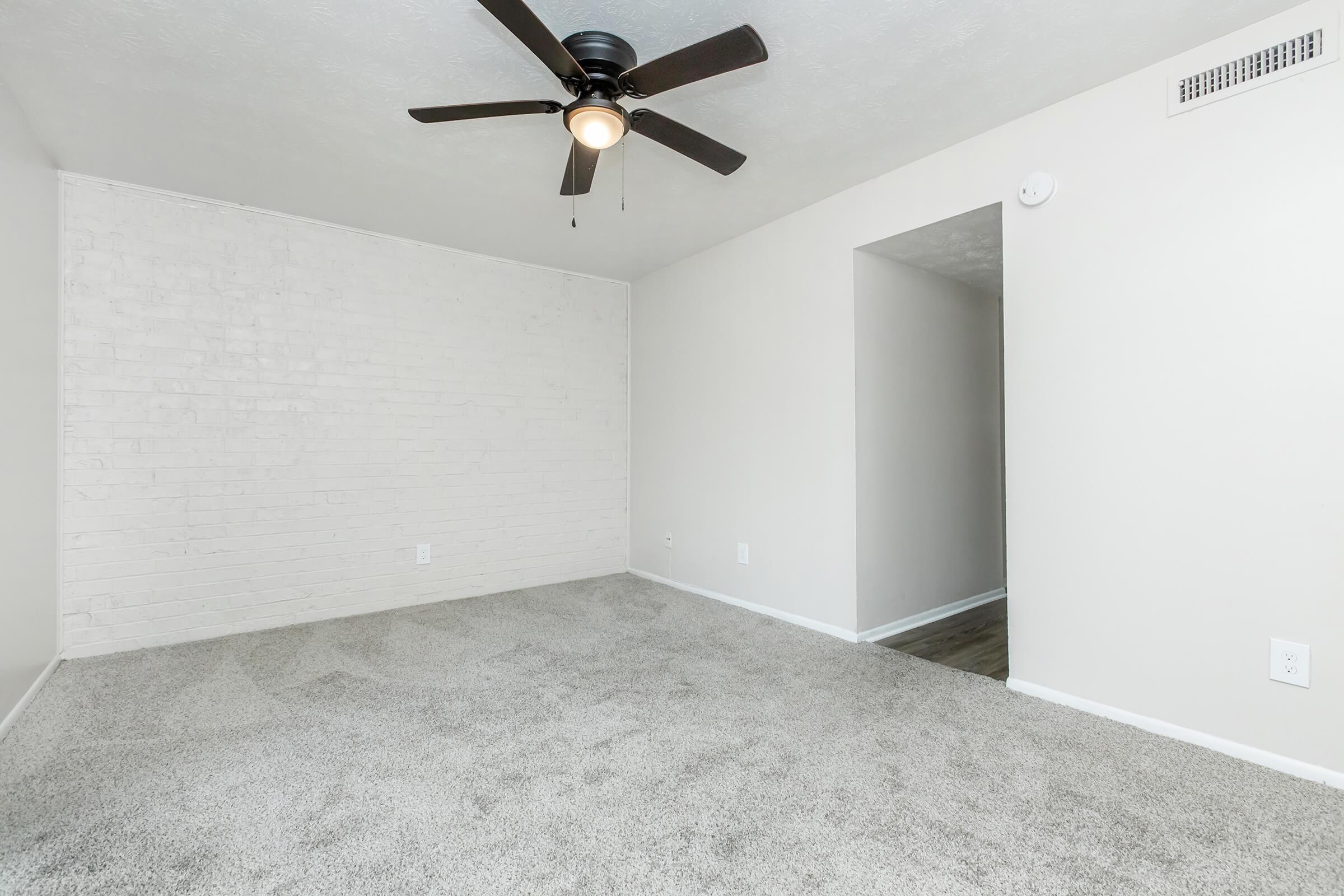 A spacious, empty room with light-colored walls and a white brick accent wall. The floor is covered in soft, light gray carpet. A ceiling fan with a light fixture is installed in the center, and there is an entryway leading to another area visible on the right side. Natural light creates a bright atmosphere.