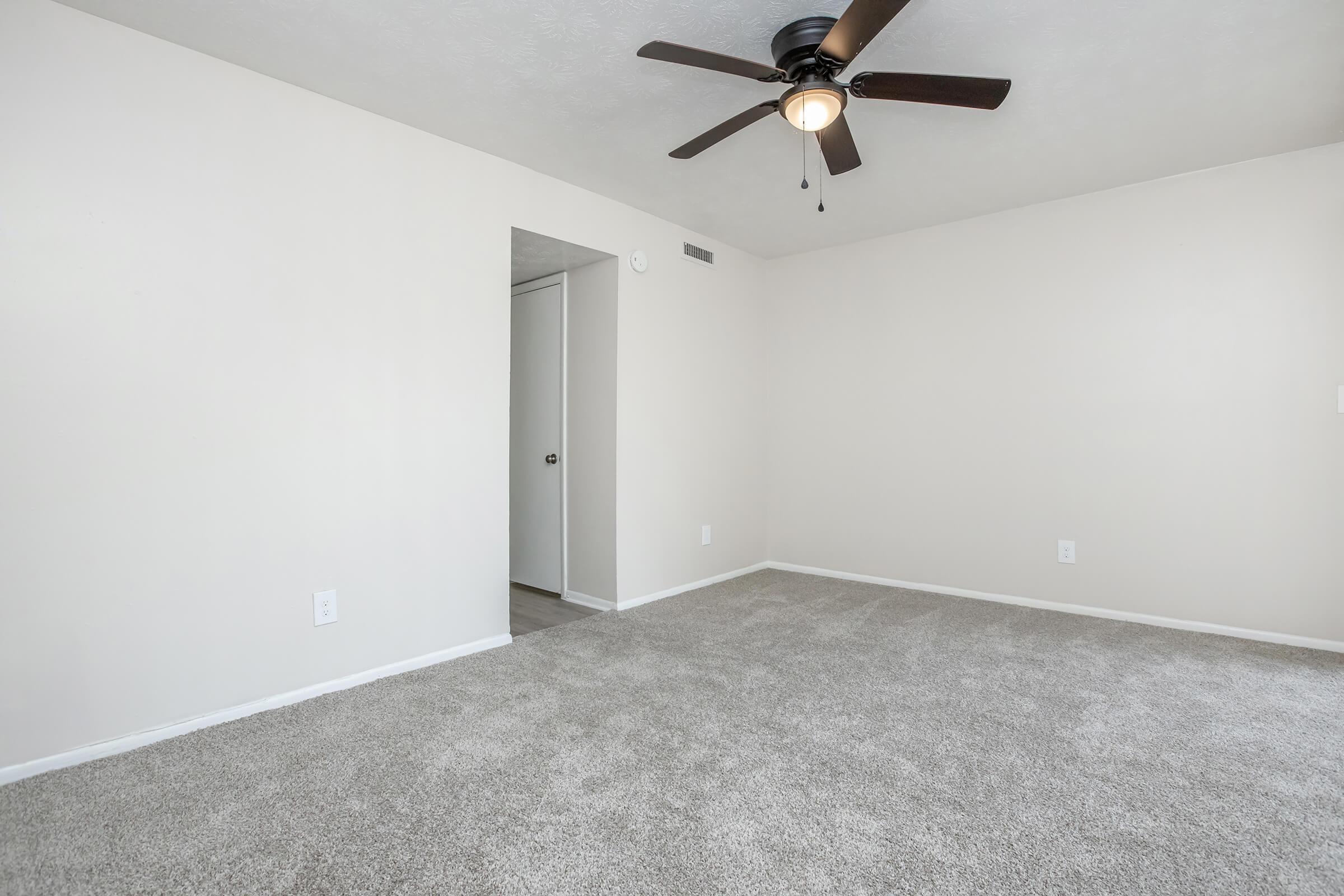 A spacious, empty room with light beige walls and a ceiling fan. The floor is covered with light gray carpeting, and there is a closed door leading to another space. The overall atmosphere is bright and open, ideal for decorating or furnishing according to personal taste.