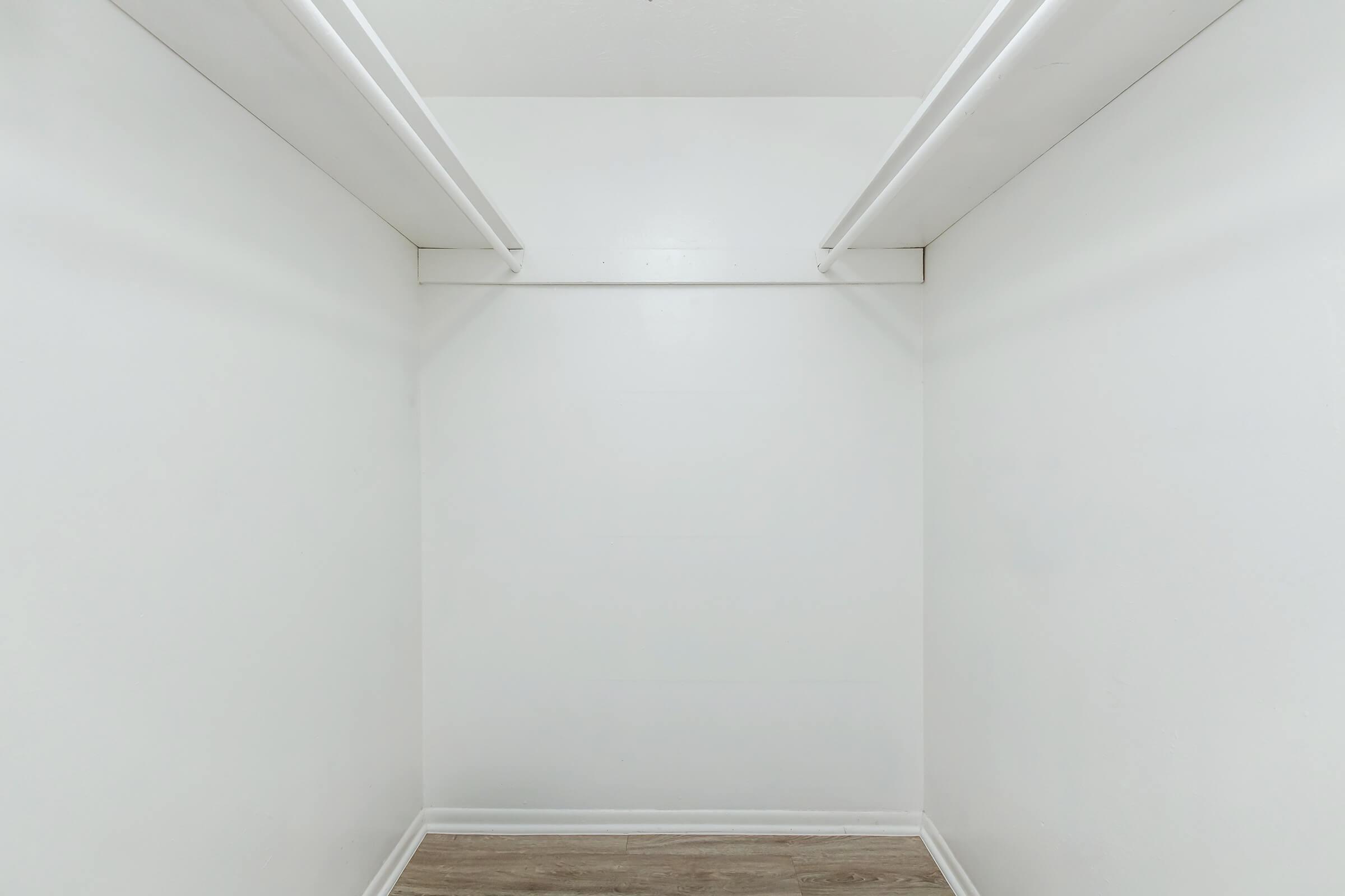 An empty closet with white walls, two horizontal shelves, and a wooden floor, creating a clean and spacious appearance.