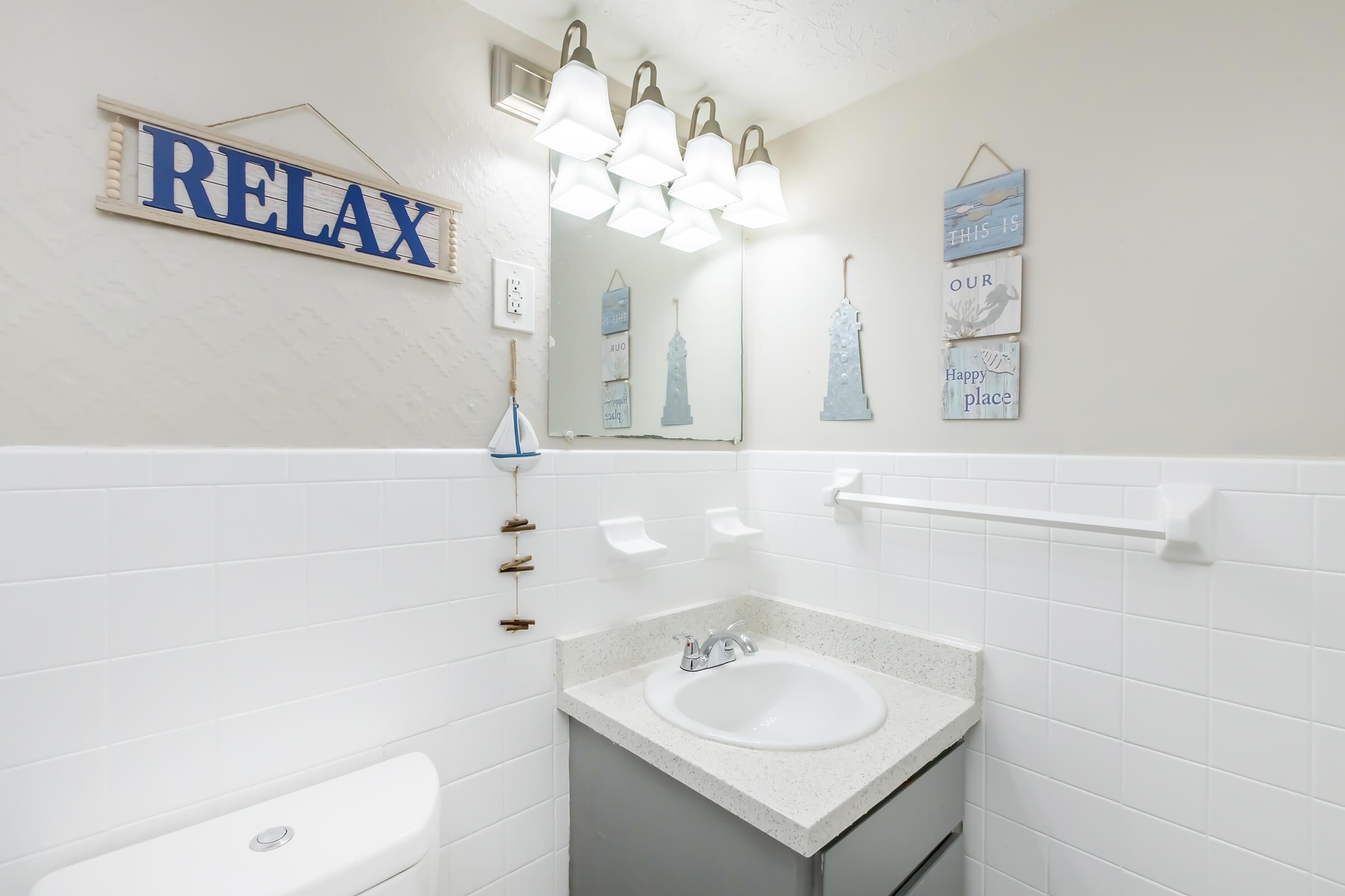 Bright and airy bathroom featuring a light-colored countertop sink with a modern faucet. Above, there are decorative wall signs with the word "RELAX" and beach-themed decor. Soft lighting is provided by four hanging light fixtures. The walls are tiled in white, creating a clean and serene atmosphere.