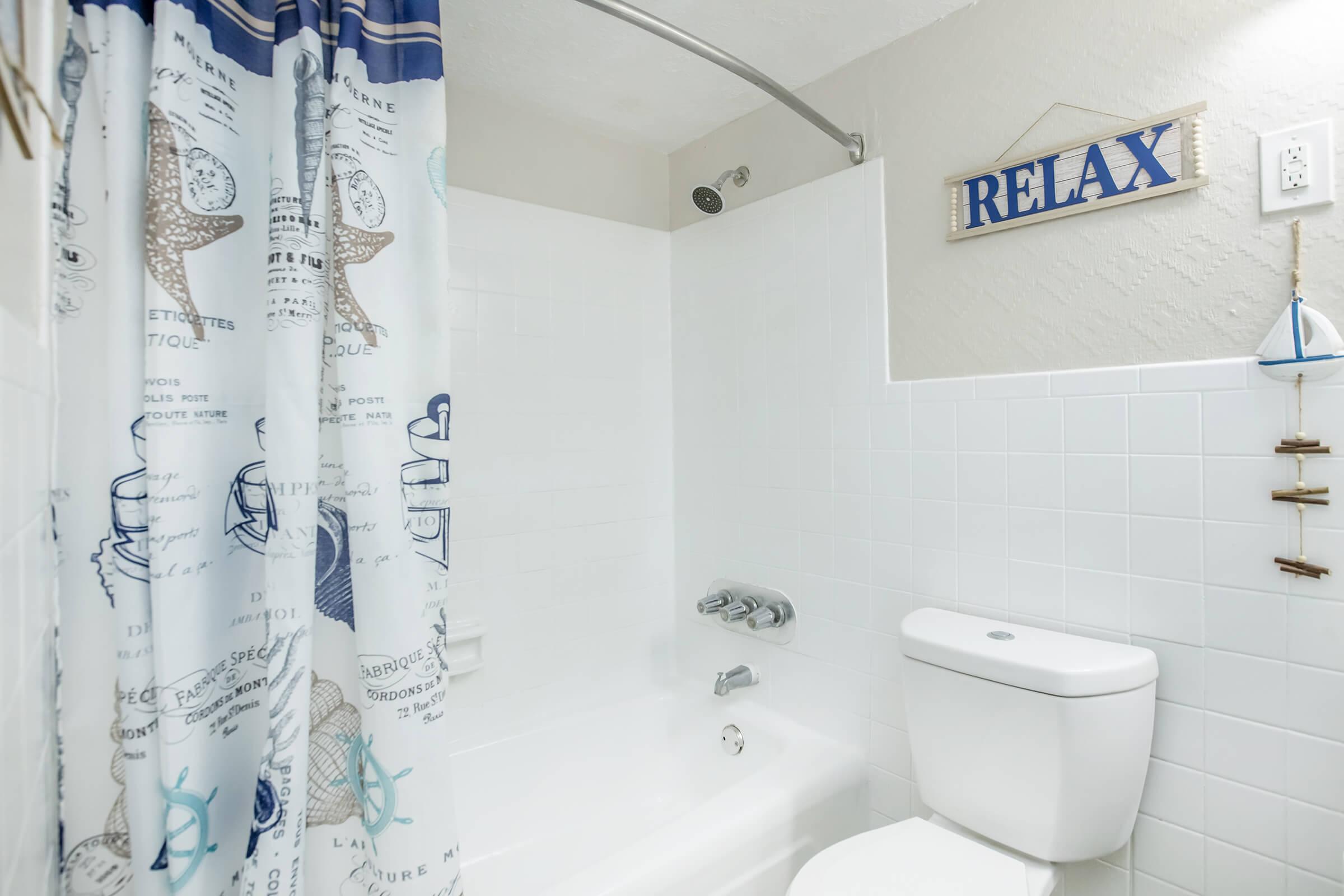 A clean and modern bathroom featuring a white bathtub, toilet, and a decorative shower curtain with a beach theme. A sign that reads "RELAX" is mounted on the wall, and a small nautical decoration hangs nearby, creating a serene and inviting atmosphere.
