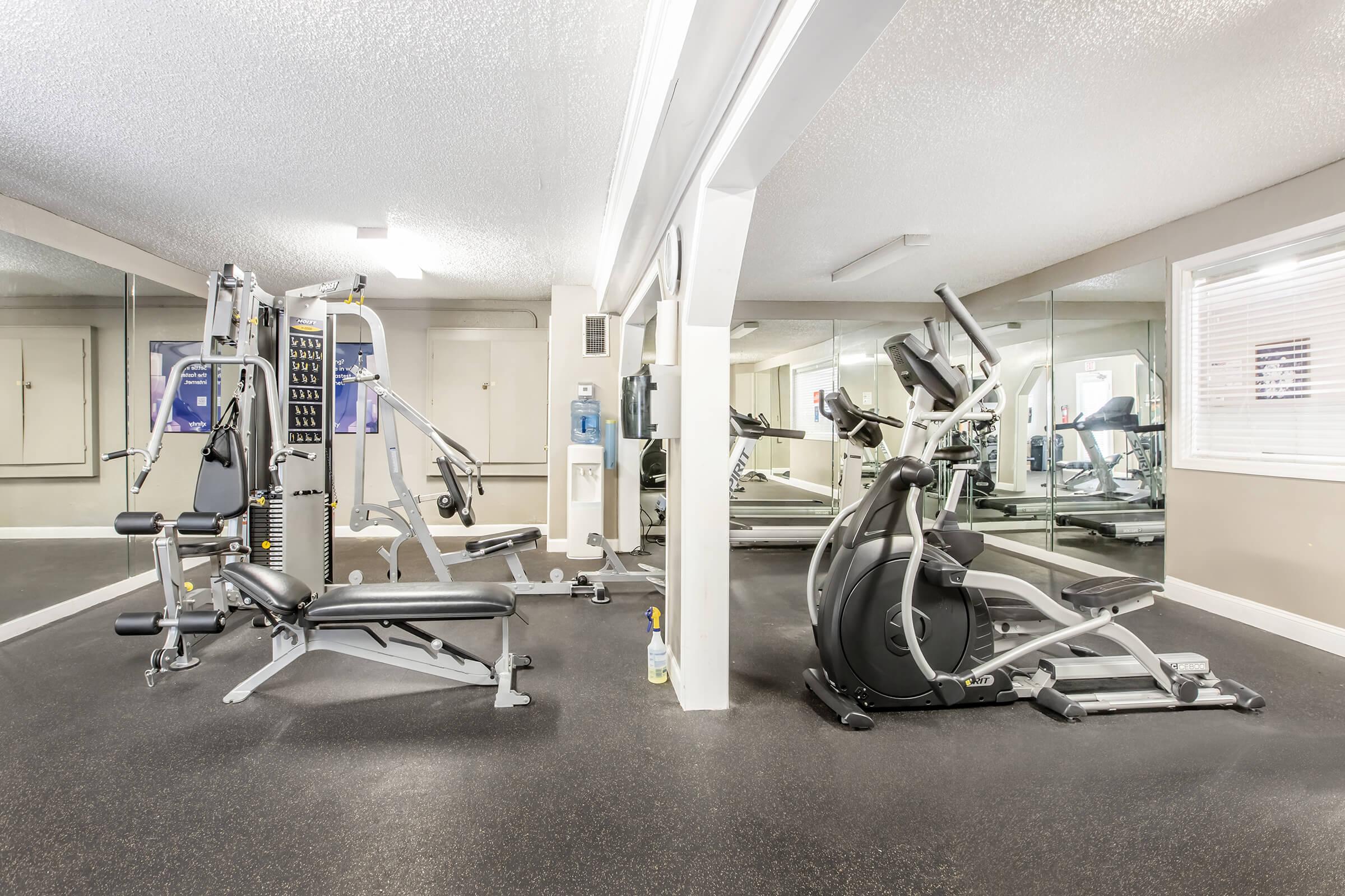 A spacious gym area featuring various fitness equipment, including a multi-station weight machine, a stationary exercise bike, and an elliptical machine. Mirrors line the walls, enhancing the sense of openness and allowing users to check their form during workouts.