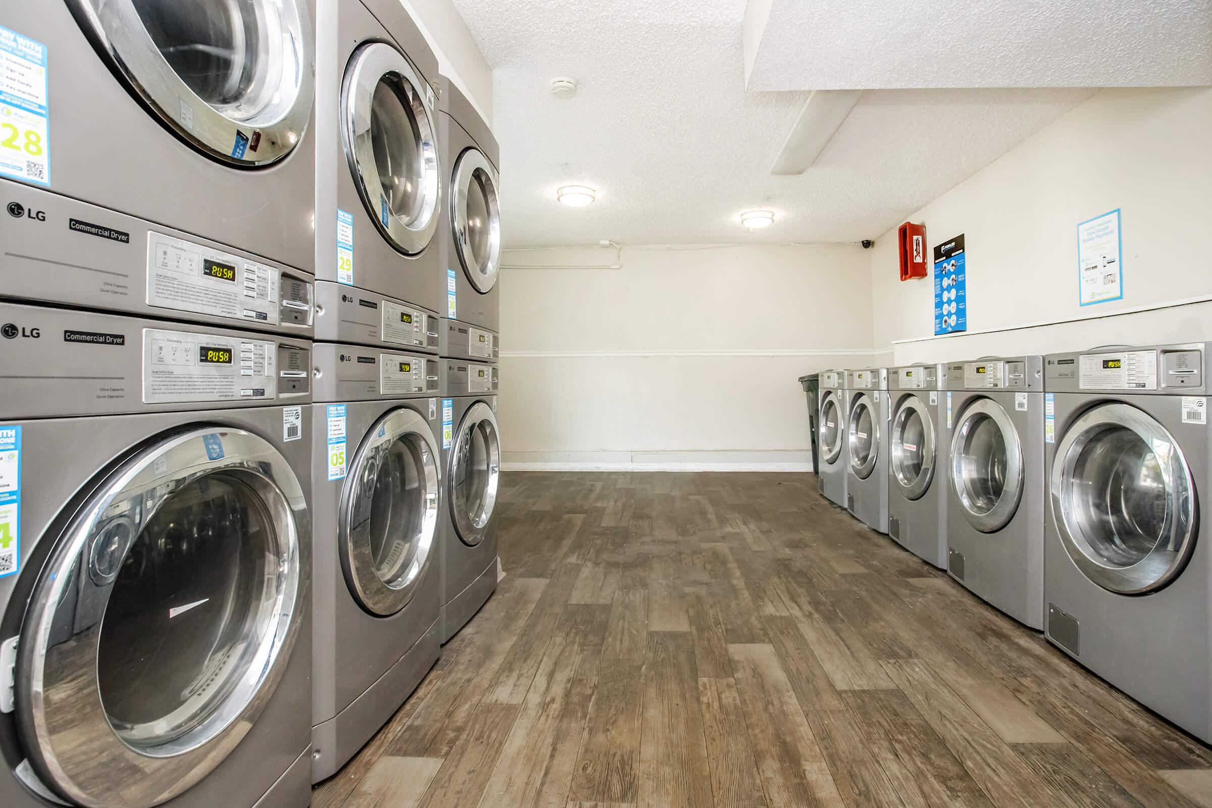 A clean and well-lit laundry room featuring several stacked silver washing machines and dryers arranged in two rows. The floor has a wood-like finish, and the walls are painted light colors, creating a modern and inviting atmosphere.
