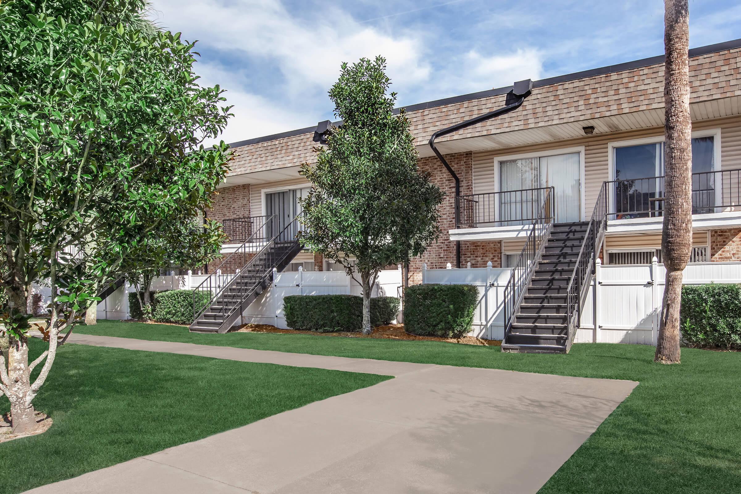 Two-story apartment building with brick exterior and balconies. There are well-maintained shrubs and small trees in front, a concrete pathway leading to the entrance, and a green lawn surrounding the area. The sky is clear with some clouds.