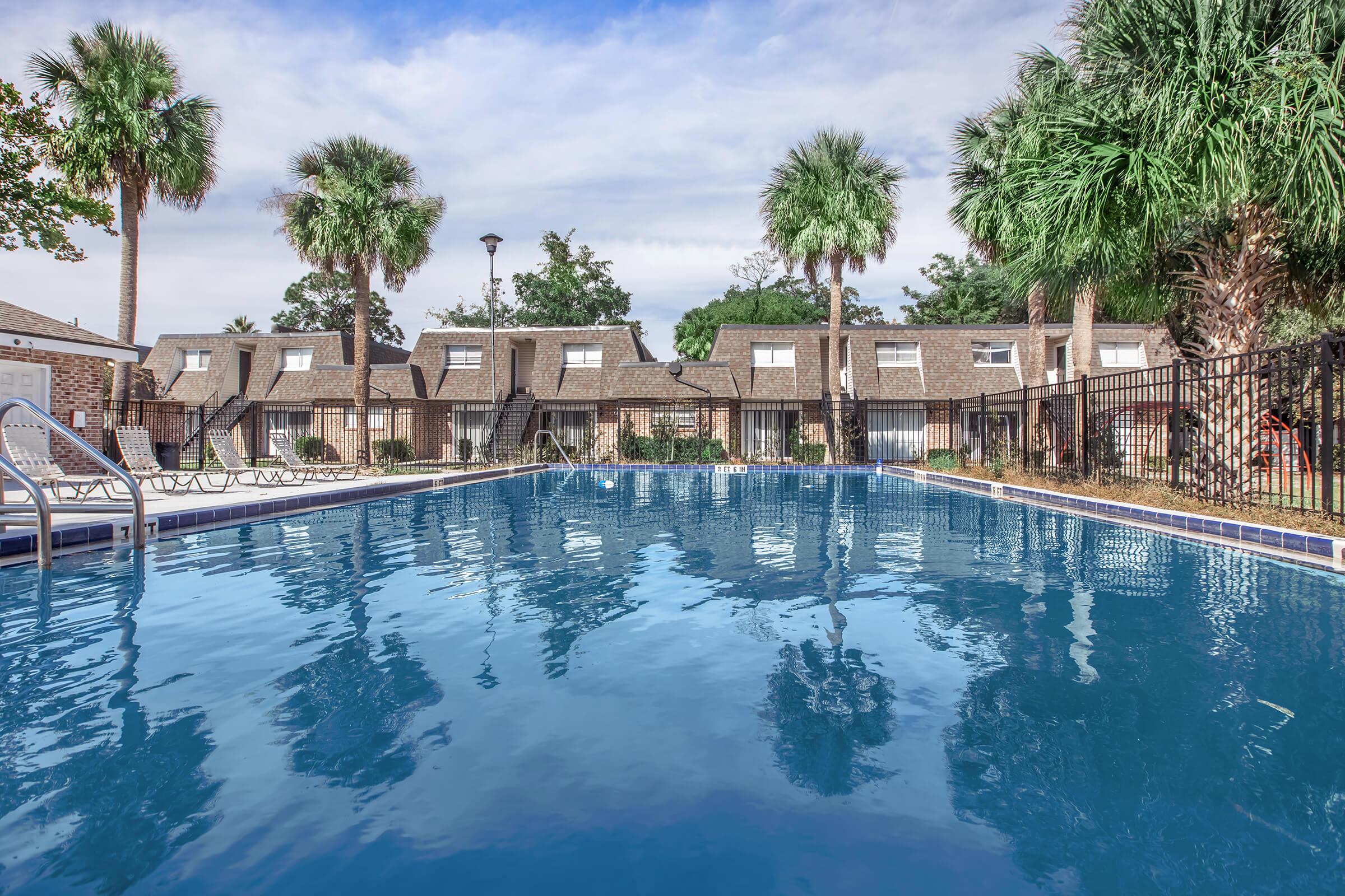 A tranquil swimming pool surrounded by palm trees, with a clear blue sky reflecting off the water. The pool area is enclosed with a fence, and in the background, there are several brown brick townhouses with peaked roofs. The scene conveys a relaxed, inviting atmosphere.