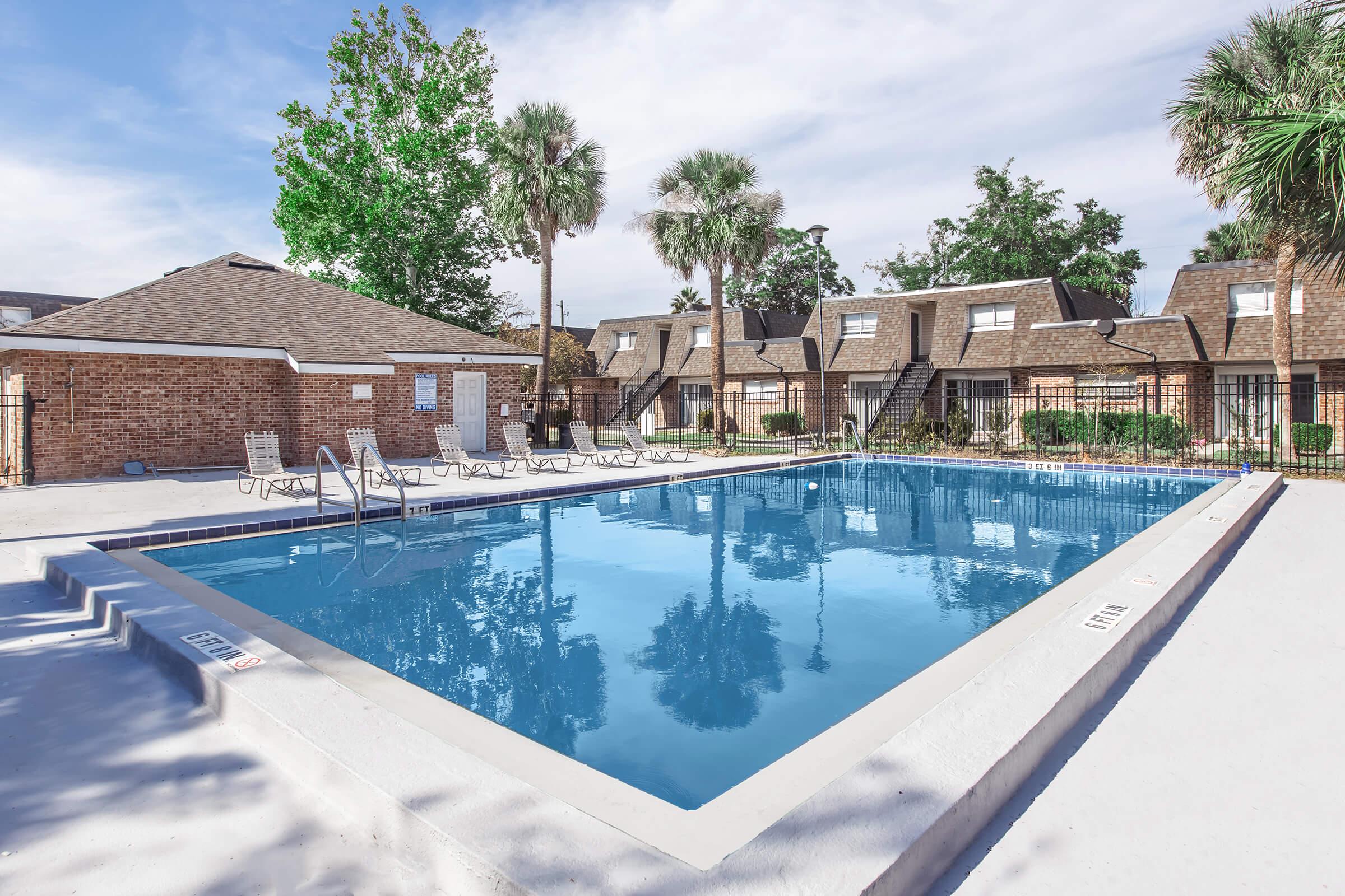 A clear, inviting swimming pool surrounded by lounge chairs and lush green trees. In the background, there are brown brick buildings with multiple stories and stairways leading to individual units, all set in a well-maintained outdoor area.