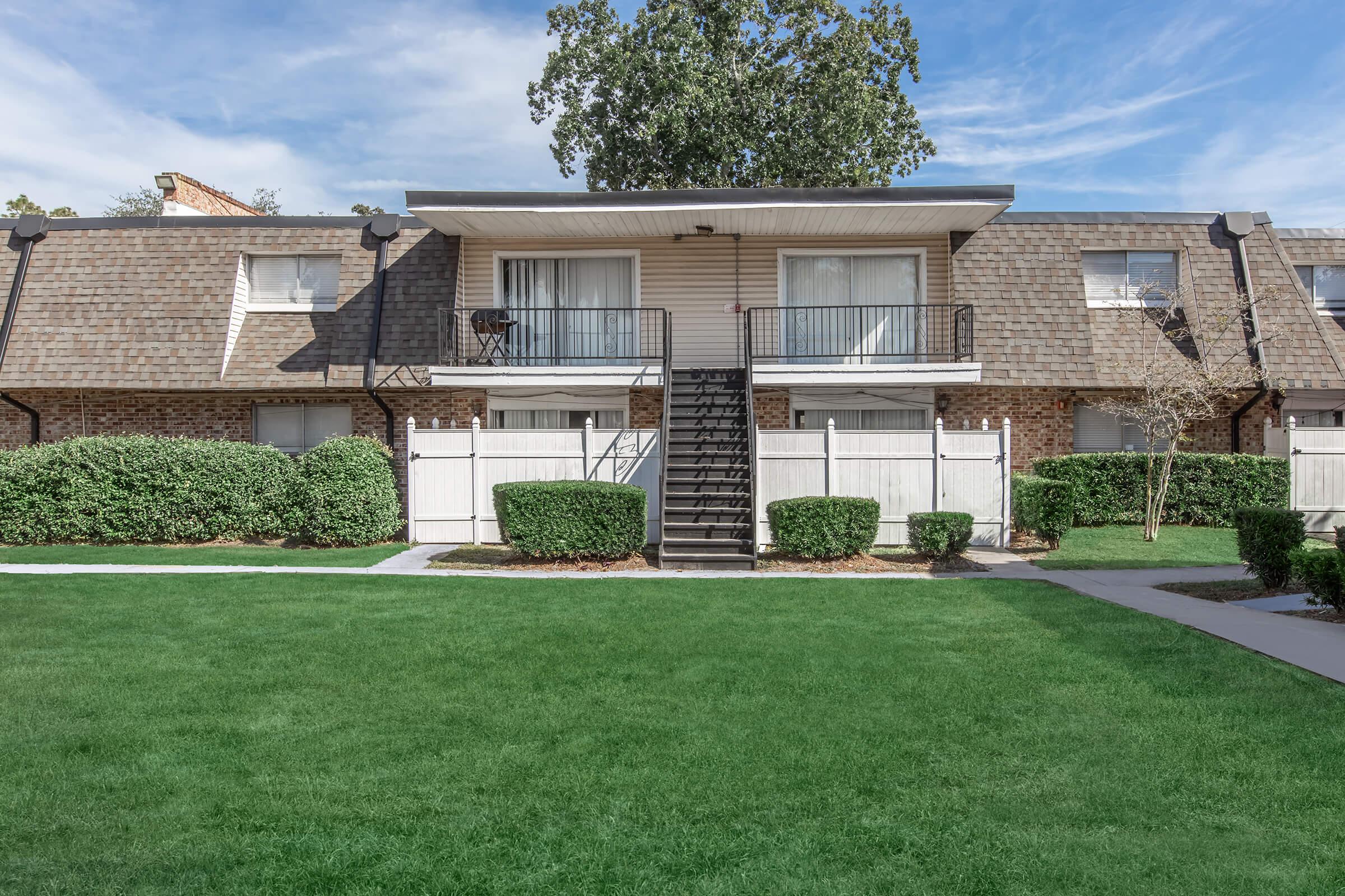 A two-story apartment building with a brown shingle roof and white balconies. The building is flanked by well-manicured green shrubs and grass in the foreground. There are two sets of stairs leading to the upper units, and a clear blue sky is visible above.