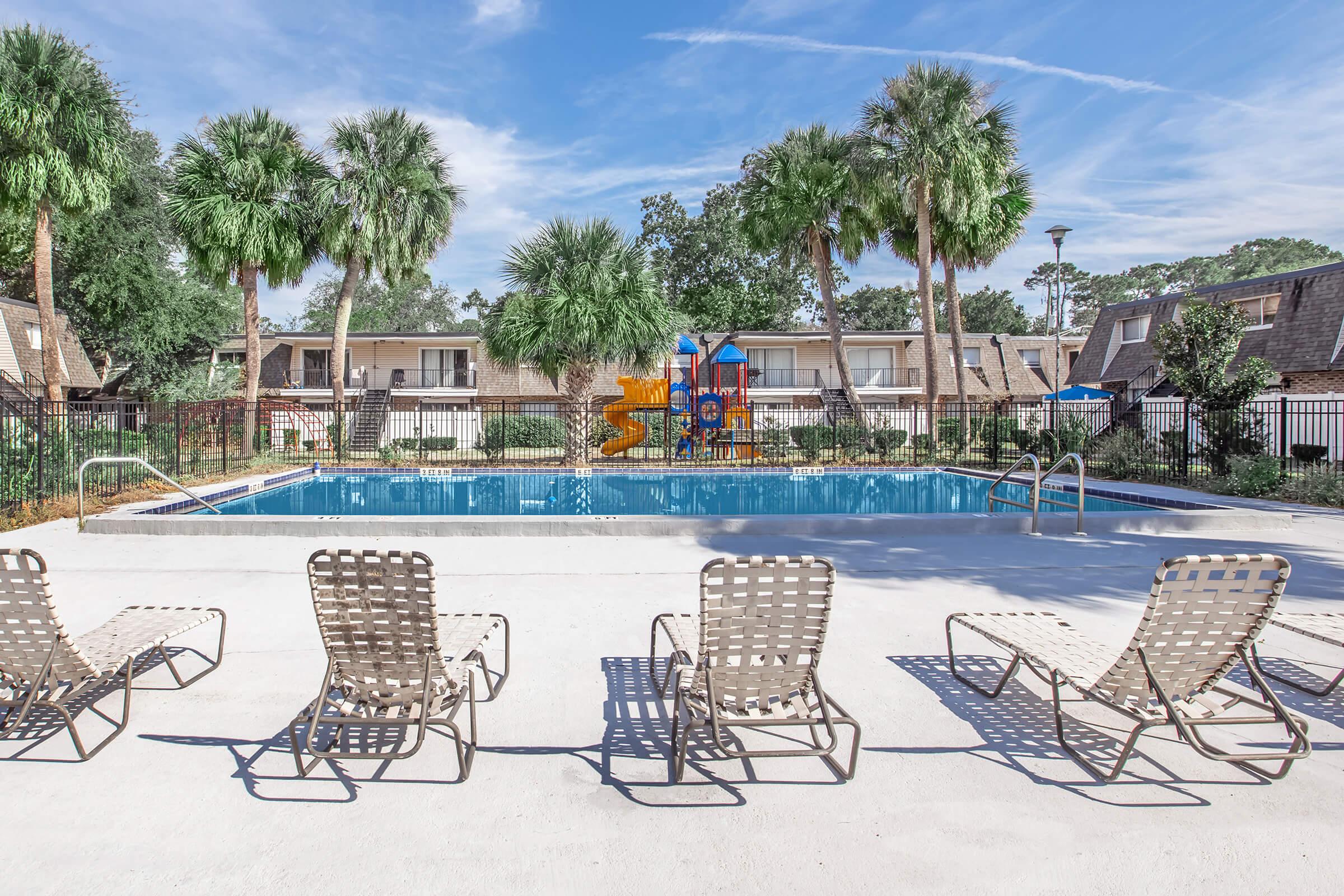 A clear blue swimming pool surrounded by palm trees and lounge chairs. At one end of the pool, there is a colorful water play structure. The background features two-story apartment buildings and a sunny sky with scattered clouds.