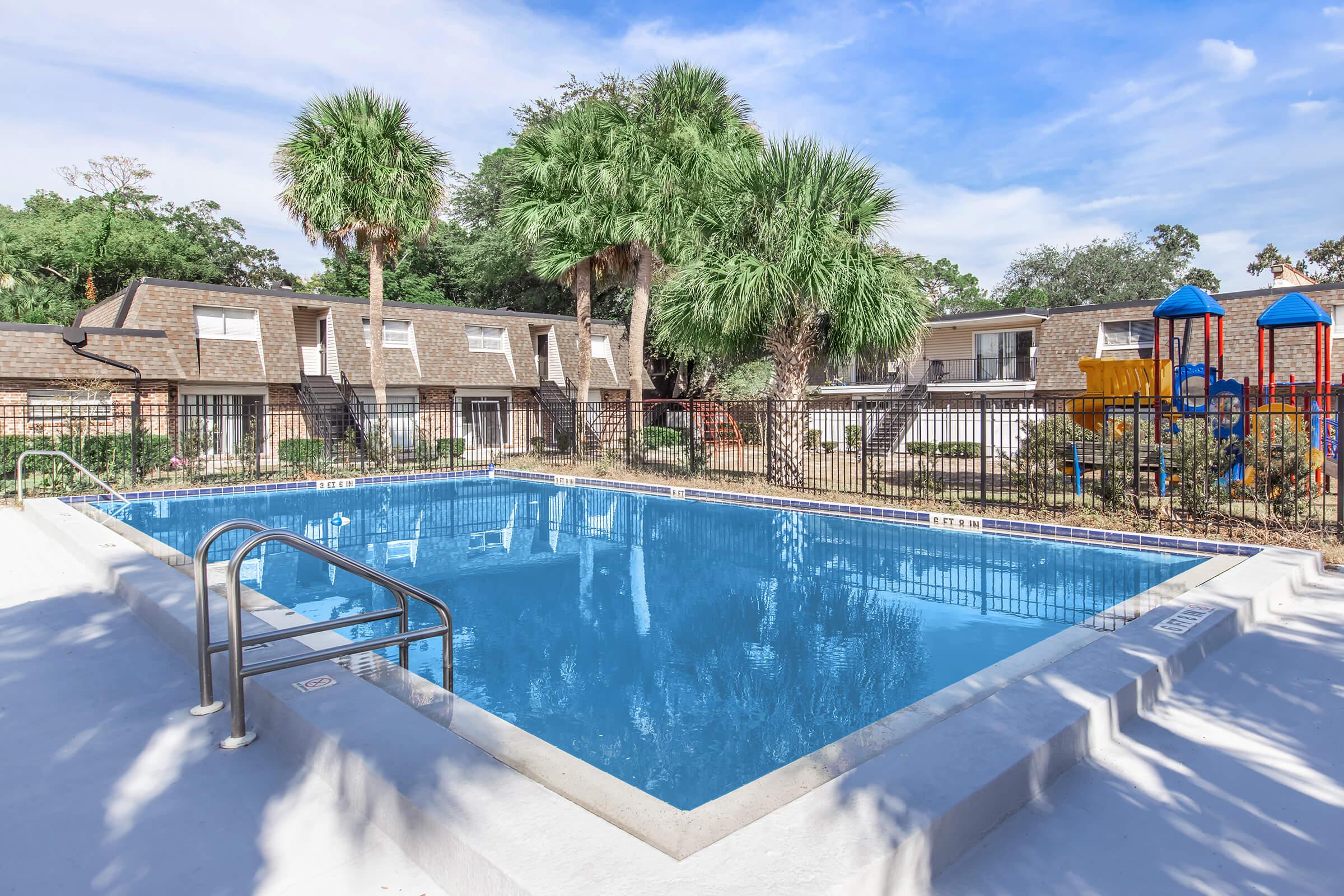 A sparkling blue swimming pool surrounded by palm trees and a black metal fence. In the background, there are multiple apartment buildings with a play area featuring colorful playground equipment. The sky is clear with a few clouds, creating a bright and inviting atmosphere.
