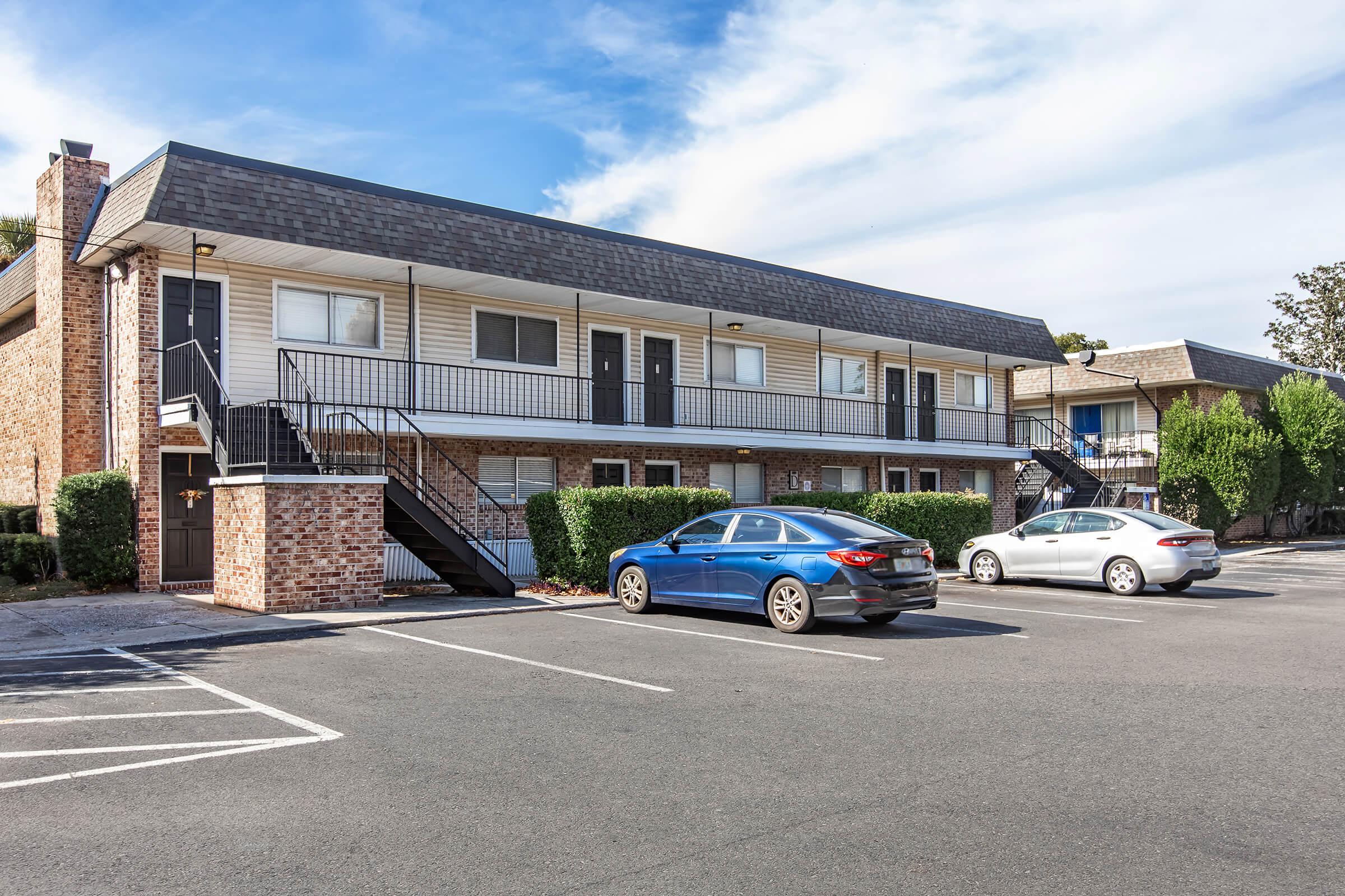 A two-story apartment building with a brick facade and multiple entrances. The building features black railings and is surrounded by neatly trimmed hedges. In the foreground, there are several parking spaces with two cars parked, one blue and one silver. The sky is partly cloudy.