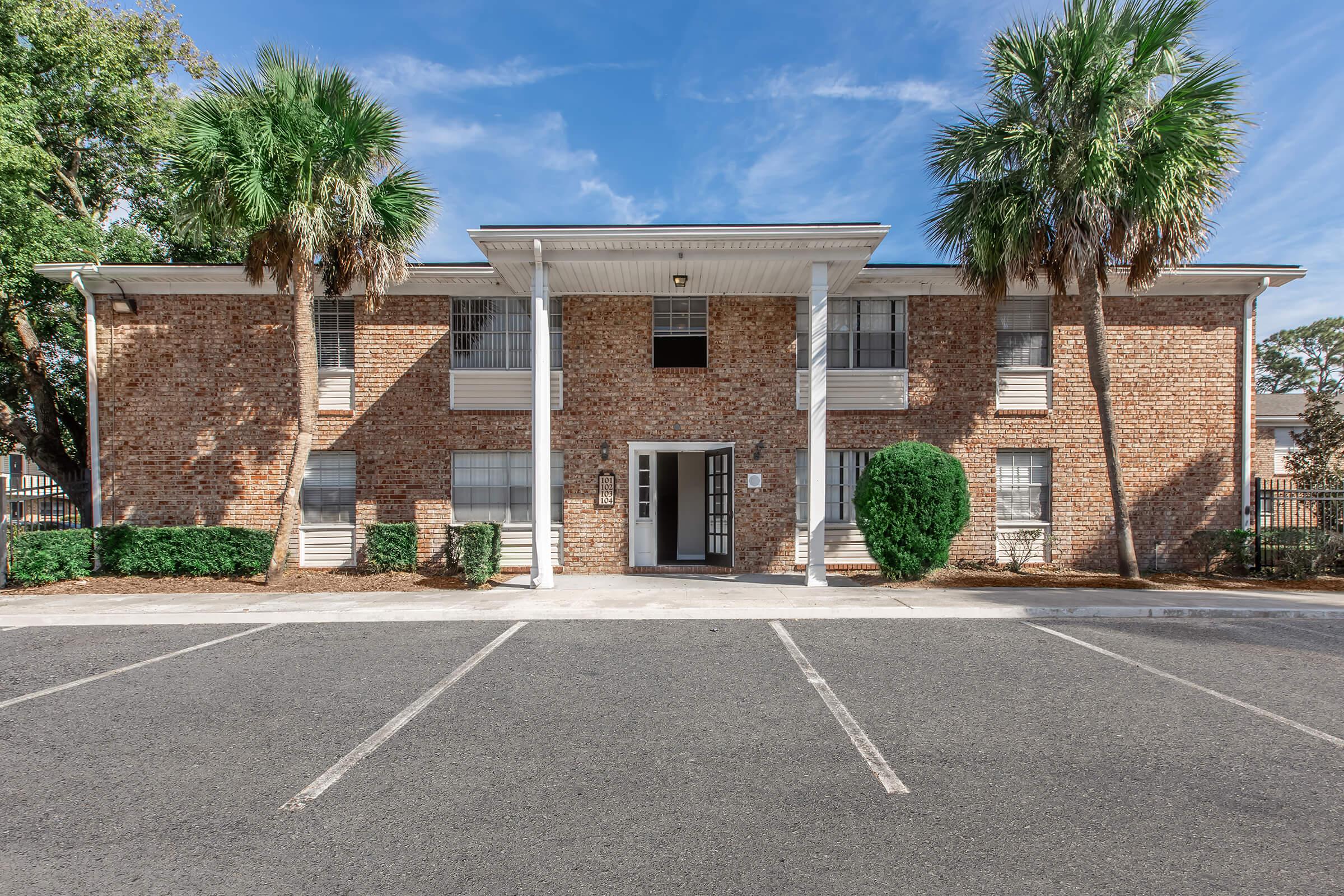 A brick apartment building with a centered entrance surrounded by palm trees. The building features multiple windows and a small landscaped area in front. There are several parking spaces visible in the foreground, and the sky is clear with a few clouds.