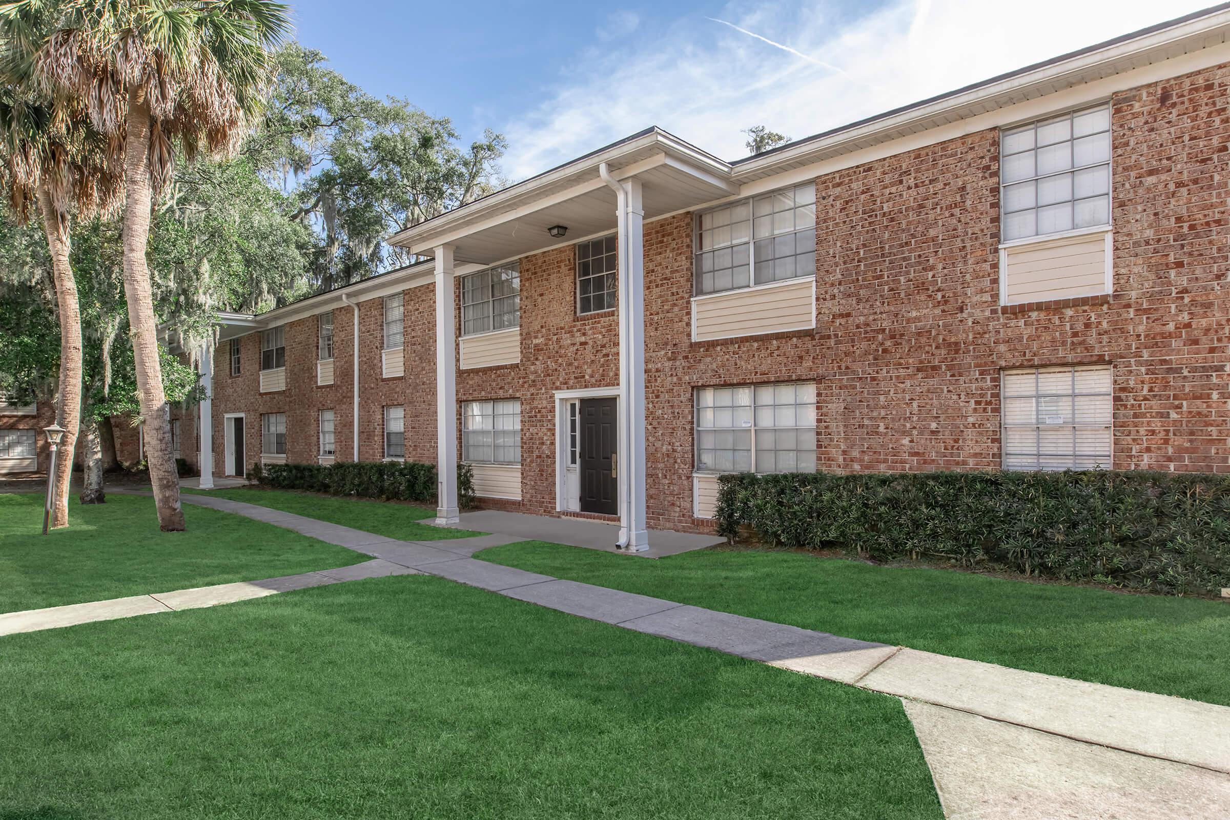 Two-story brick apartment building with white trim, featuring multiple windows and a central entrance. The building is surrounded by well-maintained grassy areas and lined with small bushes. Palm trees are visible on the left side, contributing to a sunny, inviting atmosphere.