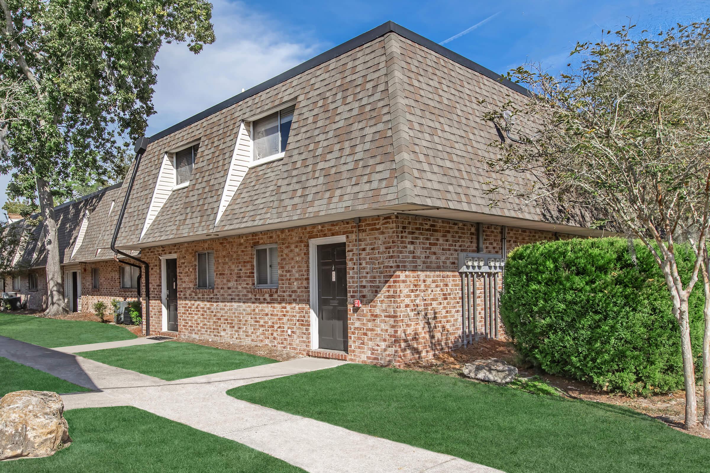 A landscaped area featuring a brick building with a steeply sloped roof and multiple windows. The building is surrounded by well-maintained grass and trees, with a pathway leading to the entrance. Bright blue sky and a few clouds are visible in the background.
